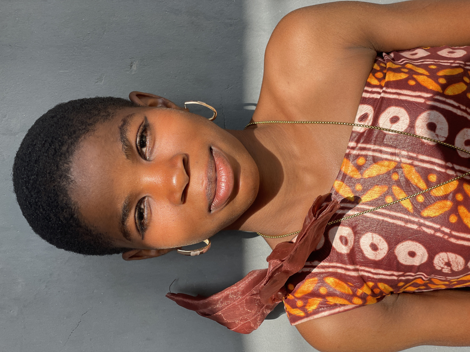 A close-up, high-angle portrait features a young person, likely female, with dark skin and short, dark, curly hair, lying down on a textured grey surface. She is looking directly at the camera with a gentle, soft smile.
The subject is adorned with large, golden hoop earrings and a thin gold beaded chain, which appears to be a body chain or waist beads, draped across her torso. She is partially covered by a vibrant, patterned fabric in shades of brown, orange, and off-white, featuring circular and abstract designs, possibly a traditional batik print. A piece of sheer, ruffled reddish-brown fabric is also visible near her shoulder.
The scene is bathed in strong, natural sunlight, casting distinct shadows on the grey background and highlighting the contours of her face and body. The light suggests a bright, sunny day, consistent with an outdoor setting in a warm climate like Badagry, Nigeria. Her expression conveys a sense of calm and ease. The grey background is plain and unadorned, appearing to be concrete or a similarly textured wall or ground. There is no visible text in the image.