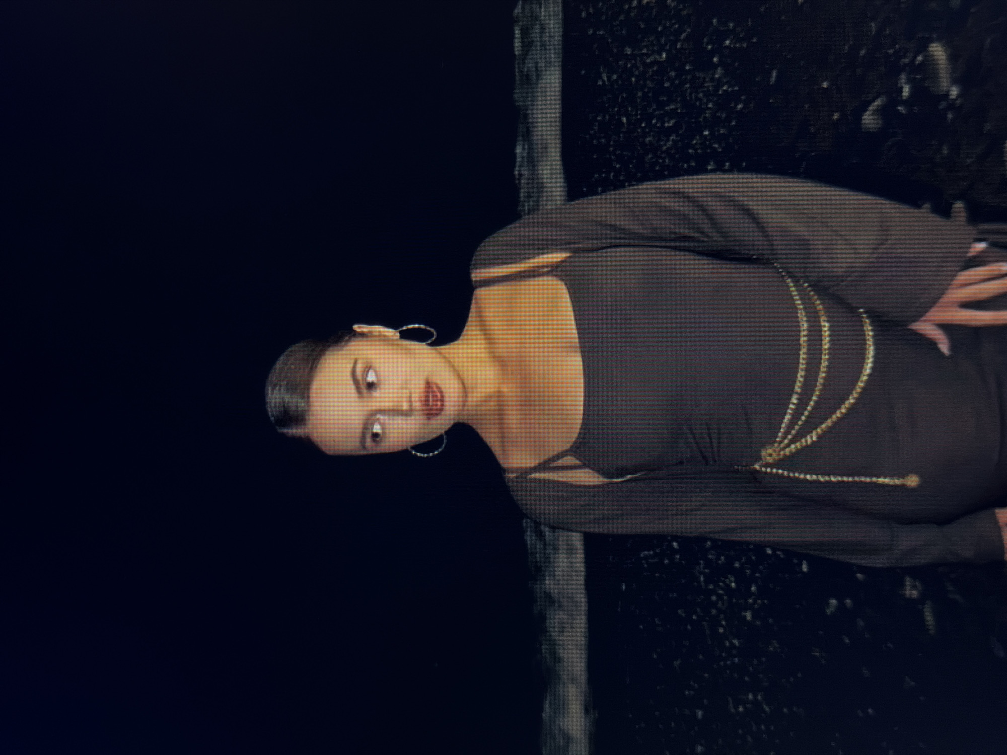 A young woman stands on a dark, pebble-strewn beach at night, facing right and looking away from the camera. She has her hair pulled back and wears large hoop earrings. Her dress is dark and appears to be made of a thin, possibly sheer material, with spaghetti straps. A layered gold chain belt is cinched around her waist. The background consists of dark water with small waves and a very dark sky. The image appears to be taken in Sirius, Russia, by the sea.
