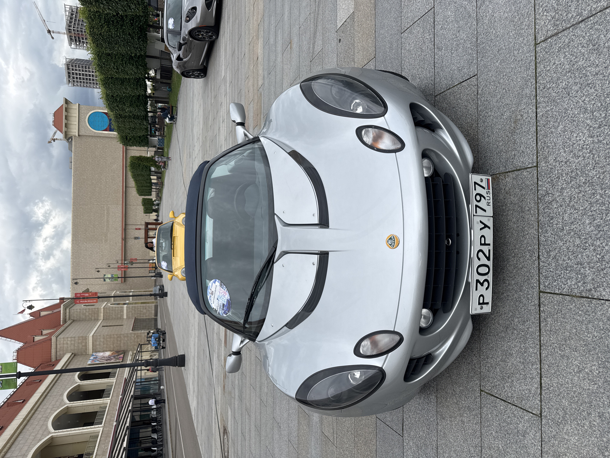 A silver Lotus Elise convertible is parked on a paved plaza in front of a building with a red roof and arched windows, possibly a shopping center or entertainment venue, in Moscow, Russia. The car's license plate reads "P302PY 797 RUS". In the background, a yellow convertible is parked further back, and a gray Alfa Romeo sports car is visible to the right. The environment appears to be an urban setting with trees lining the plaza and construction cranes visible in the distance. The sky is overcast.
