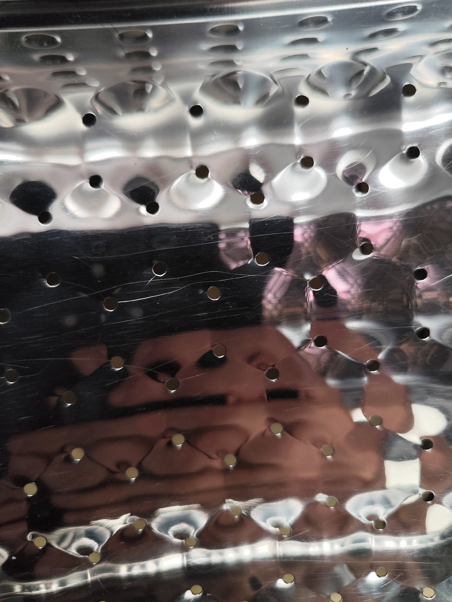 This media file is a close-up, abstract shot of a metallic surface with a pattern of circular indentations and small holes. The surface is highly reflective, showing distorted shapes and colors from its surroundings.
The primary subject is the textured metallic material itself, likely part of an appliance like a washing machine drum, characterized by its dimpled, perforated design. There are no discernible people or animals as the main subjects, although blurred reflections of what appear to be human figures and possibly fabric are visible on the metallic surface. The scene is indoors, given the reflections and the sterile nature of the metallic surface. There are no clear time of day or weather cues.
Notable details include numerous scratches on the metallic surface, indicating wear and use. The reflections are complex, showing a mix of darker, shadowed areas and brighter, almost white highlights, suggesting a light source is present but not directly visible. The overall impression is one of texture, reflection, and a slightly abstract industrial aesthetic. No text is visible in the image. The context of Almaty, Kazakhstan, does not provide specific visual cues within the image itself.