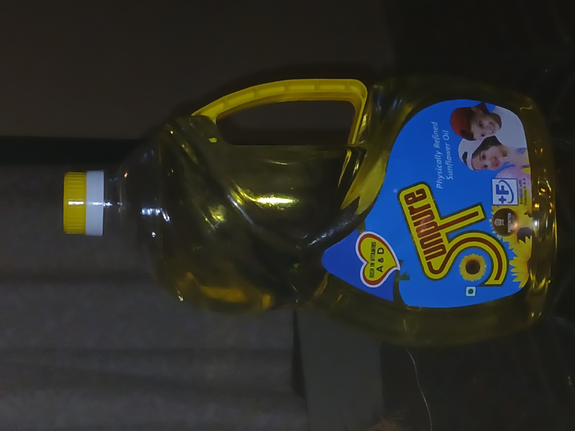 The image shows a plastic bottle of SUnpure Sunflower Oil. The bottle is yellow and has a blue label. The label has the words "SUnpure" and "Physically Refined Sunflower Oil" on it. There are also pictures of two children on the label. The bottle has a yellow handle and a white cap with a yellow band. The background is dark and out of focus. The location in the city of Bengaluru, India cannot be inferred from the image.