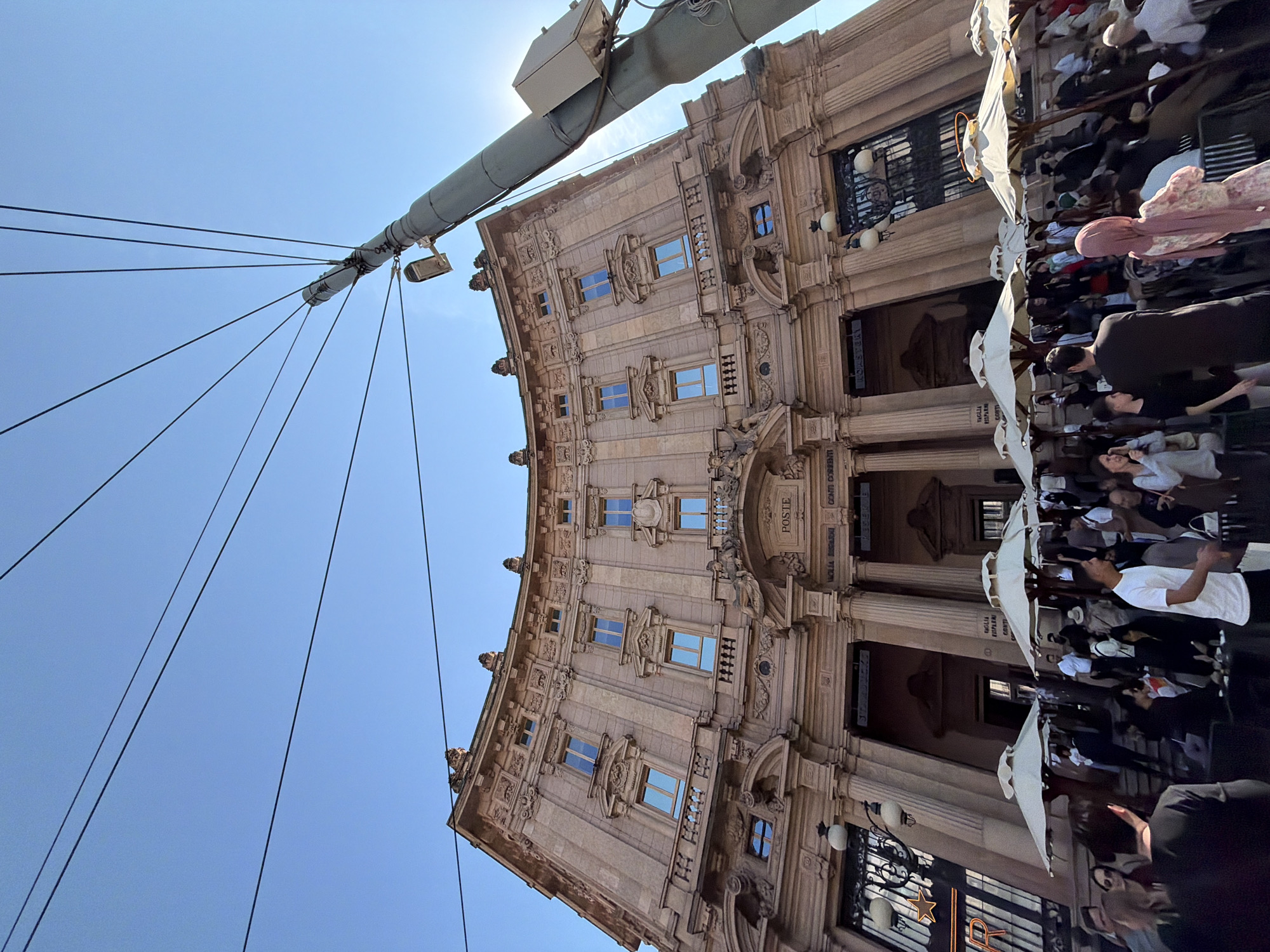 The image shows the Piazza Cordusio in Milan, Italy, with the historic Palazzo delle Poste (Post Office Palace) as the primary architectural feature. The view is from a low angle, looking upwards and slightly tilted.
The Palazzo delle Poste is a large, ornate building with a light-colored facade. It has several floors with multiple windows. A sign with the word "POSTE" is visible on the building.
In the foreground, there's a tall, gray metal pole with multiple wires extending from it.
To the right of the building, there are a number of people. Some are standing, while others are seated under white umbrellas. This suggests an outdoor café or seating area. The people appear to be casually dressed, engaging in typical tourist activities.
The sky is clear and blue. The overall impression is that of a bright, sunny day in a busy urban setting.
