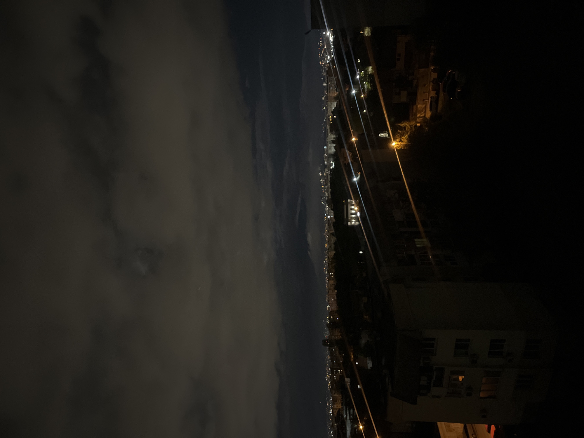 The image shows a night view of a city, likely Sofia, Bulgaria, with a dark, cloudy sky on the left. City lights stretch along the horizon, indicating buildings and infrastructure. In the foreground on the right, there is a building with visible windows and streetlights. The image appears to be taken from a high vantage point. The scene is dark, and the primary light sources are artificial lights from the city and buildings.