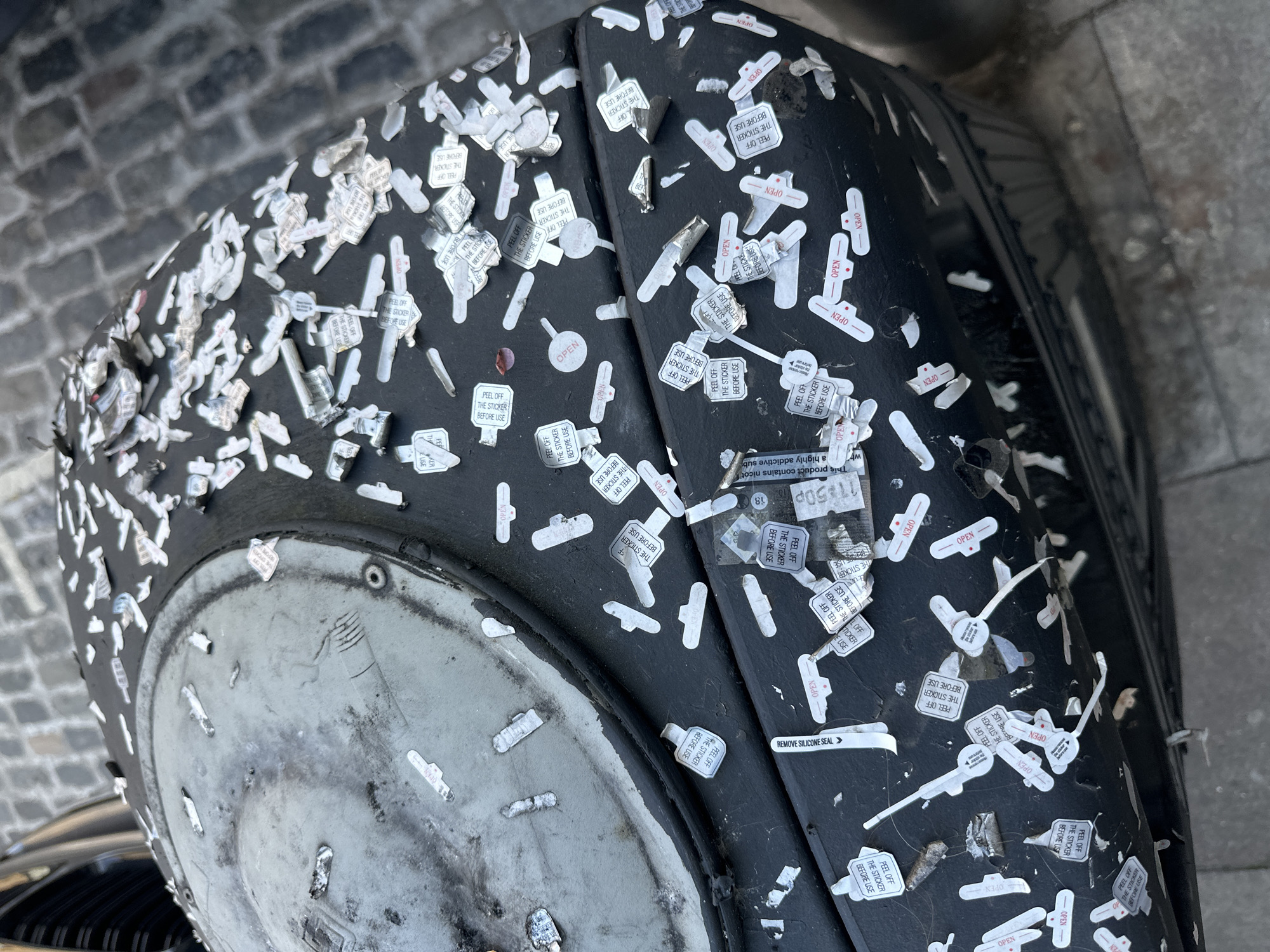 An overhead shot captures a heavily stickered, dark, cylindrical object, likely a public waste bin or planter, located on a street in London, United Kingdom. The object's dark, textured surface is almost entirely obscured by a dense accumulation of small, white stickers, many of which are peeling or partially detached. The flat, circular top of the object is a grimy, metallic grey, showing signs of wear and dirt.
No human activity is depicted; the scene is static. The stickers themselves represent a form of informal public interaction or defacement. The background features cobblestone paving on the left side, indicating an urban setting, and a darker, smoother paved surface on the right. The lighting suggests daylight, possibly overcast.
A multitude of stickers display recurring phrases such as "PEEL OFF THE STICKER BEFORE USE" and "OPEN." Other identifiable text includes "REMOVE SILICONE SEAL" and fragments from a warning label: "This product contains nicot a highly addictive sub," implying the stickers originate from tobacco or vaping product packaging. Numerical details like "11:50p" and "18" are also visible.
