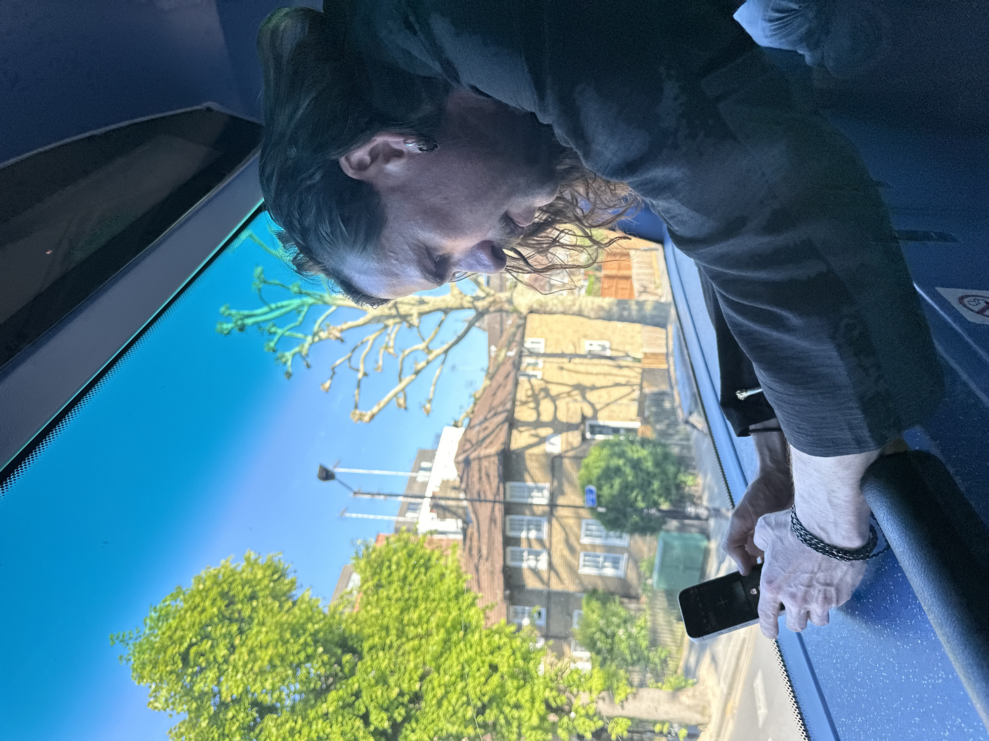 The image, taken with a 90-degree counter-clockwise rotation, captures a person inside a vehicle looking out a large window at a sunny street scene.
A male individual with long, dark, wavy hair, a beard, and a mustache is seen in profile, leaning forward from the right side of the frame. He wears a dark long-sleeved shirt or jacket, and an earring is visible in his left ear. On his right wrist, he sports a dark braided bracelet. His right hand holds a black smartphone, which rests near the window frame. His gaze is directed downwards, suggesting he is looking at his phone or something immediately outside the vehicle.
The interior of the vehicle features dark blue surfaces, possibly parts of a dashboard or window ledge, and a black armrest or handle. A red circular prohibitory sign, likely indicating "no food or drink" or "no littering," is partially visible on the blue surface to the right of the person's hand.
Outside the clear window, a vibrant streetscape unfolds under a bright, clear blue sky. Multi-story buildings with traditional architecture, featuring numerous windows and what appears to be brickwork, line the street. Lush green trees with full foliage are prominent, indicating either spring or summer. Strong sunlight casts clear shadows from the trees and buildings. A street lamp can also be seen.
The bright conditions suggest it is a clear day, likely morning or afternoon. Given the provided context, the scene is set in London, United Kingdom, viewed from what appears to be public transport, such as a bus.