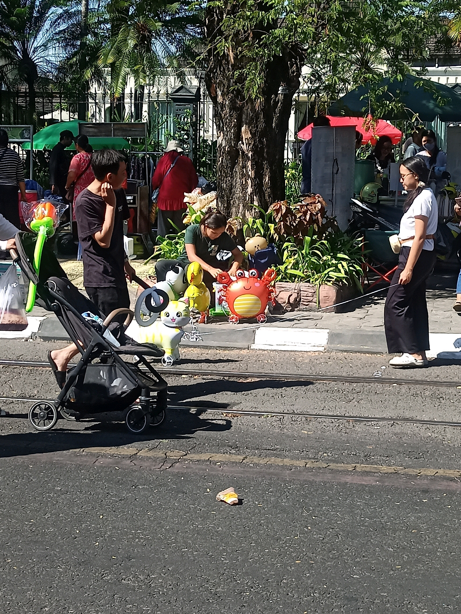 An eye-level, medium shot captures a street scene in Surakarta, Indonesia, under a bright, sunny sky. In the foreground, a black stroller sits on a cracked asphalt road, with two parallel tram tracks running across it. A person's legs, clad in black flip-flops, are visible near the stroller. The stroller contains a white and yellow cat-shaped balloon and a black steering wheel. To the right of the stroller, a red crab-shaped balloon is tethered to the ground.
Behind the stroller, a man in a black t-shirt and dark pants stands with his hand on his chin, appearing to observe the surroundings. He is facing left. To his right, a person wearing a green t-shirt is crouched down, interacting with balloons. Further to the right, a woman in a white t-shirt and black skirt stands facing left, holding a white cord that is connected to the red crab balloon. She has a cream-colored pouch slung over her shoulder.
In the background, a bustling street market is set up under trees and large umbrellas. People are browsing at stalls selling various items, including clothing and food. Palm trees and other foliage are visible, along with a white building with a black fence. The overall atmosphere suggests a lively market day in the city.
