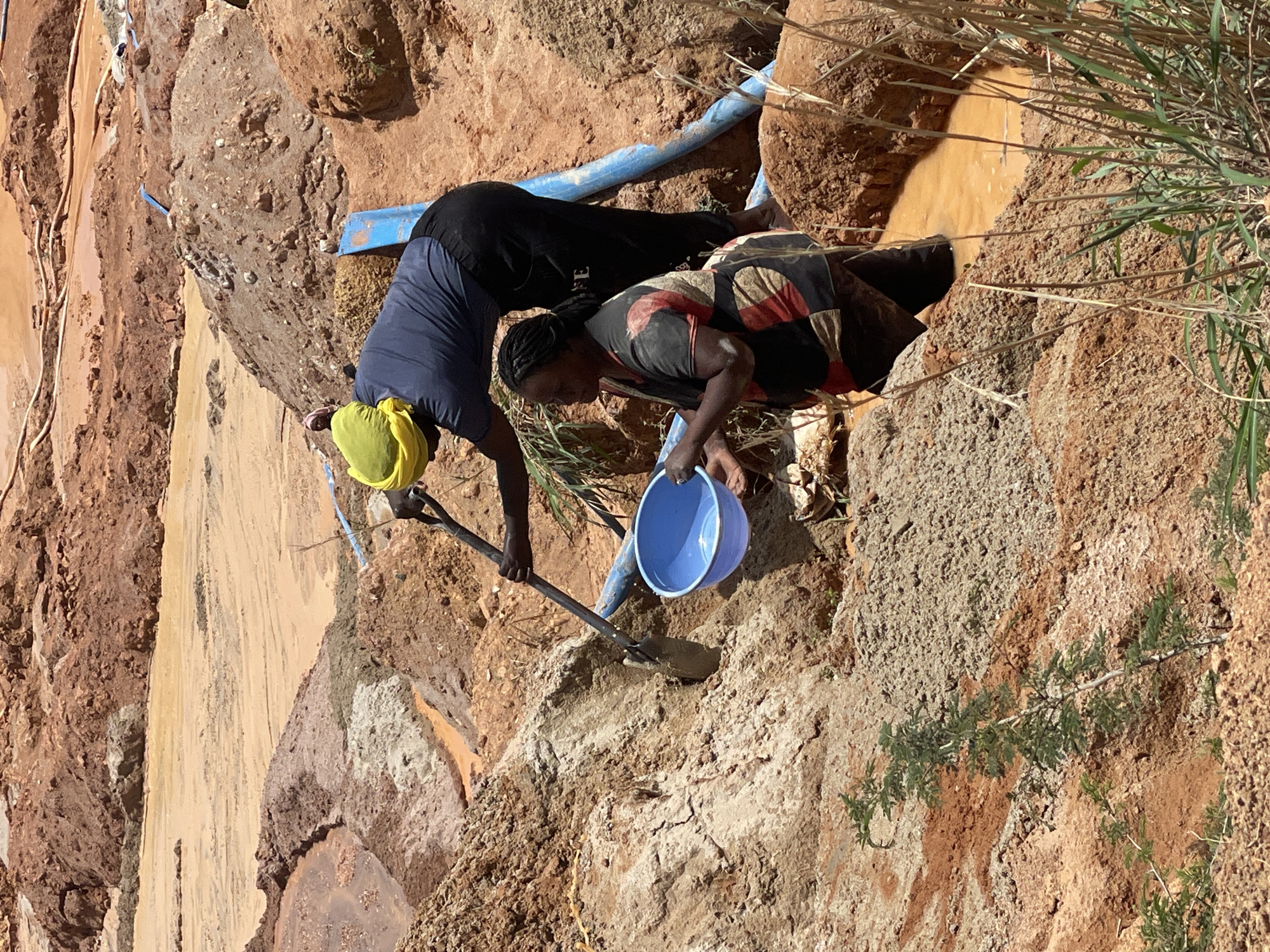 Two women are engaged in mining activities in a dry, arid environment, likely a mining site, in Jos, Nigeria. The terrain is characterized by reddish-brown soil, sand, and some exposed rocks. In the foreground, one woman, wearing a dark blue t-shirt and black pants, is using a shovel to dig into the dirt. She is also holding a blue plastic bowl in her left hand. Another woman, wearing a patterned dress with red, black, and white colors, stands partially submerged in muddy, orange water in a trench or pit. She is holding a blue bowl. Blue pipes are visible in the background, suggesting water or material transport. The overall scene depicts manual labor in a challenging, resource-extraction setting.