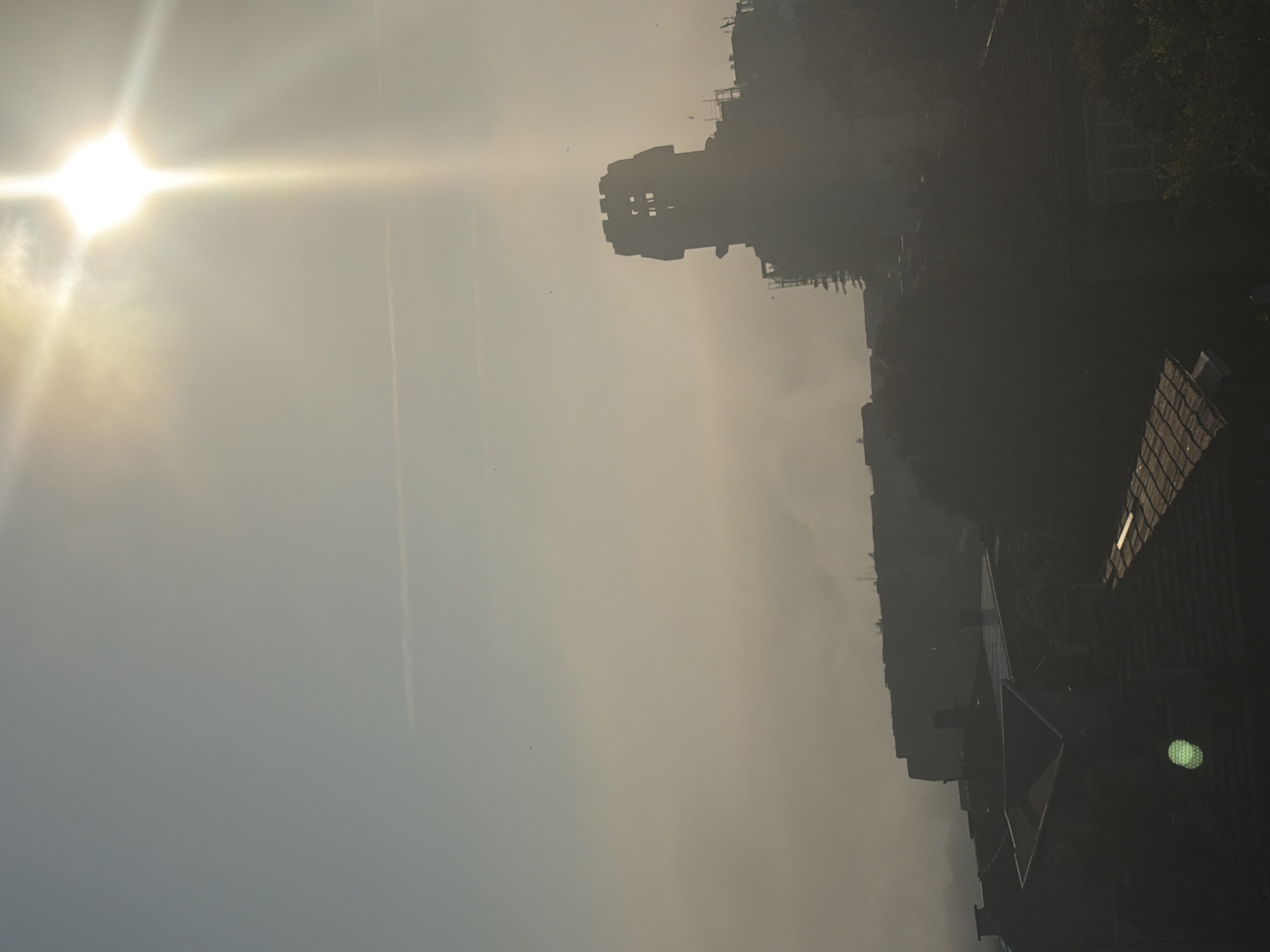 The image captures a hazy, sunlit scene, likely during early morning or late afternoon, with a strong emphasis on atmospheric light. The bright, diffused sun, positioned in the upper left when oriented correctly, dominates the sky, emitting intense light rays and exhibiting a prominent lens flare. A subtle halo or circumsolar arc is visible around the sun, indicating specific atmospheric conditions.
Against this luminous backdrop, a distinctive, dark silhouette of an old, fortified building or castle tower stands prominently in the upper right. This structure, possibly built of stone, features crenellated battlements at its top, suggesting historical architecture. Below and to the right of this main structure, other buildings and rooftops are visible as dark silhouettes, their forms partially obscured by the ambient haze. Some rooftops show visible texture, contrasting with the smooth silhouettes of other elements.
Small, indistinct specks, possibly birds, are scattered across the sky, while a faint, thin linear mark, resembling a contrail, extends across the upper-mid section of the sky. The pervasive haze creates a dreamlike or ethereal quality, causing all elements to appear as dark outlines against the radiant sky.
The overall scene conveys a sense of stillness and peacefulness, with the intense, diffused sunlight providing a dramatic backdrop. Given the context of Shumen, Bulgaria, the silhouetted structure could be a part of the Shumen Fortress or another historical landmark. There are no visible people or discernible activity, and no visible text in the image.