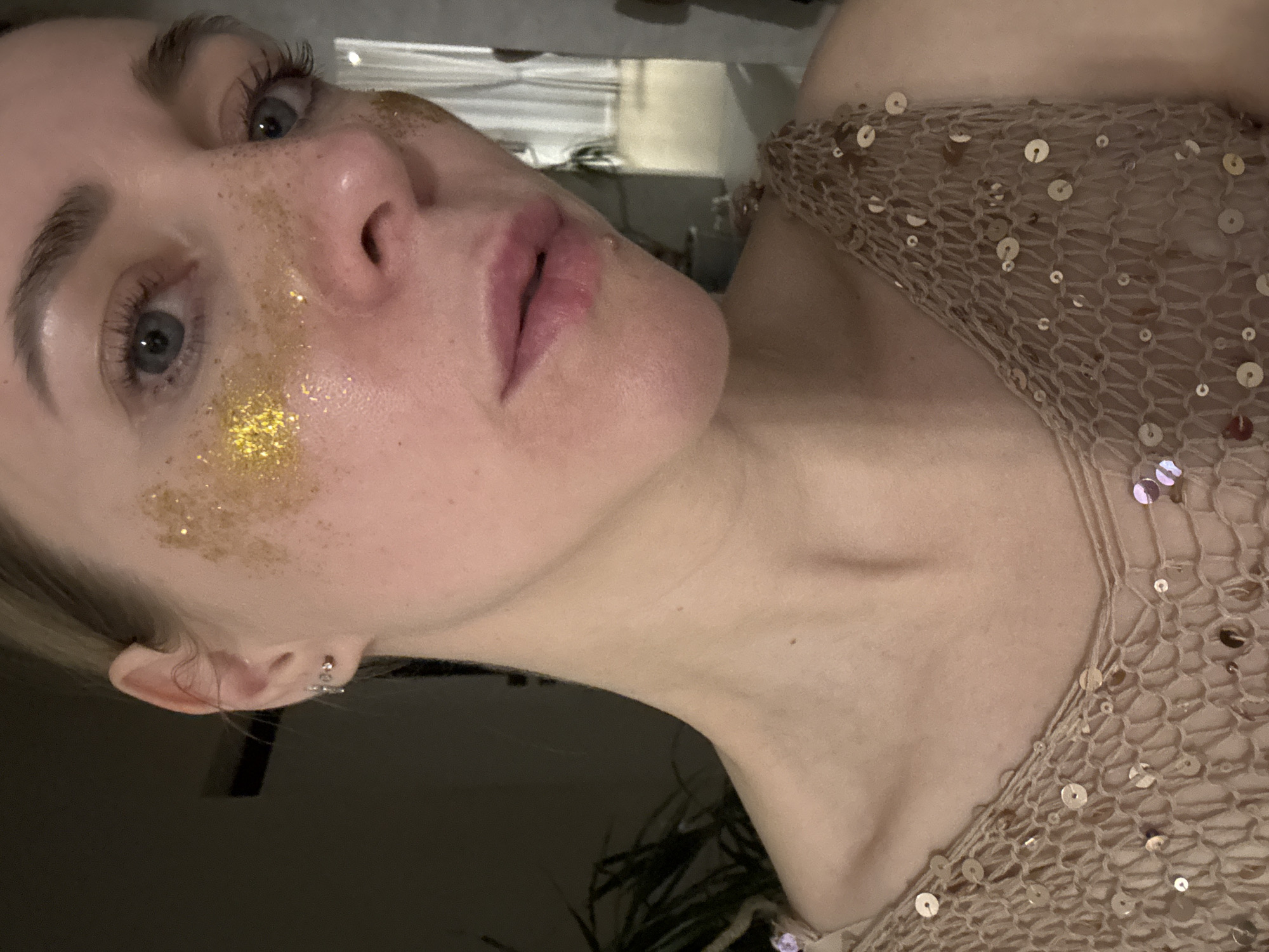 A close-up, rotated portrait features a young woman looking upwards and to the right of the frame, with her head positioned on the left side of the image and her body extending to the right. She has light-colored eyes, possibly blue or grey, and full, pinkish lips. Her fair skin is adorned with scattered golden glitter on her cheeks, giving the appearance of decorative freckles or an artistic embellishment. Her eyelashes appear long and dark.
She is wearing a light beige or tan mesh garment, possibly a top or dress, which is decorated with numerous small, shiny sequins. A small, clear earring is visible in her left earlobe, with another smaller, possibly sparkling, earring further up her ear. Light-colored hair, likely blonde, is visible near her ear. Her neck and upper chest are visible, appearing smooth.
The background is dark and indistinct, suggesting an indoor setting with low lighting, likely during the evening or night. No specific activity is depicted beyond posing for the photograph. The overall impression is one of a stylized or festive portrait. The image contains no visible text. This scene is set in Chaikovsky, Russia.