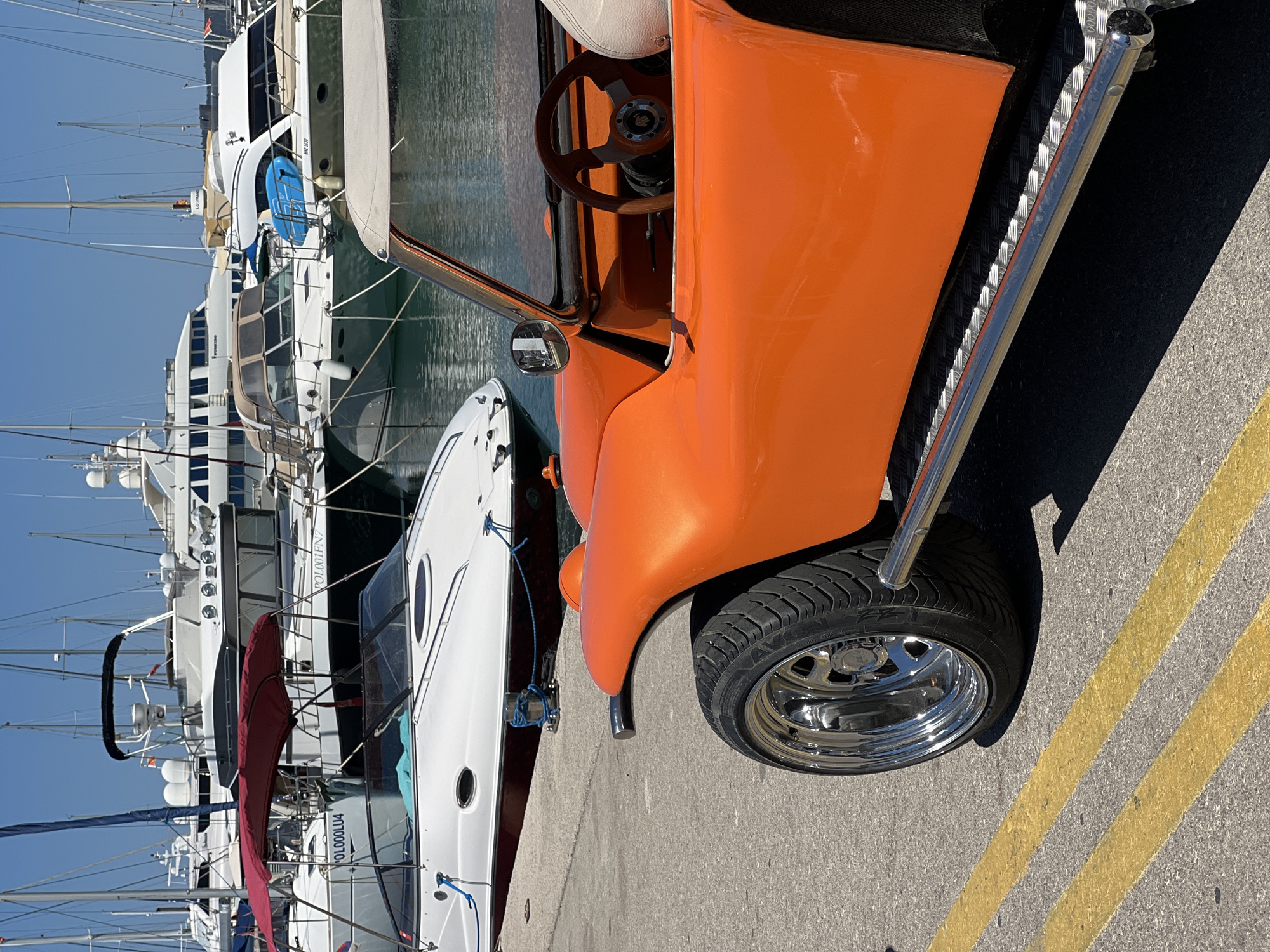 An orange dune buggy is parked on a paved area with yellow lines, in front of a harbor filled with various yachts and boats. The dune buggy is in the foreground, taking up the right side of the frame. It has a chrome bumper and a bright orange body. The interior features a wooden steering wheel and a white seat. In the background, a white motorboat with a red canopy is docked. The water of the harbor is calm and reflects the blue sky. The scene is brightly lit by the sun, indicating a clear day. The location appears to be a marina in the city of Bar, Montenegro, judging by the presence of numerous yachts and the general ambiance of a Mediterranean port. There are no people visible in the image.
