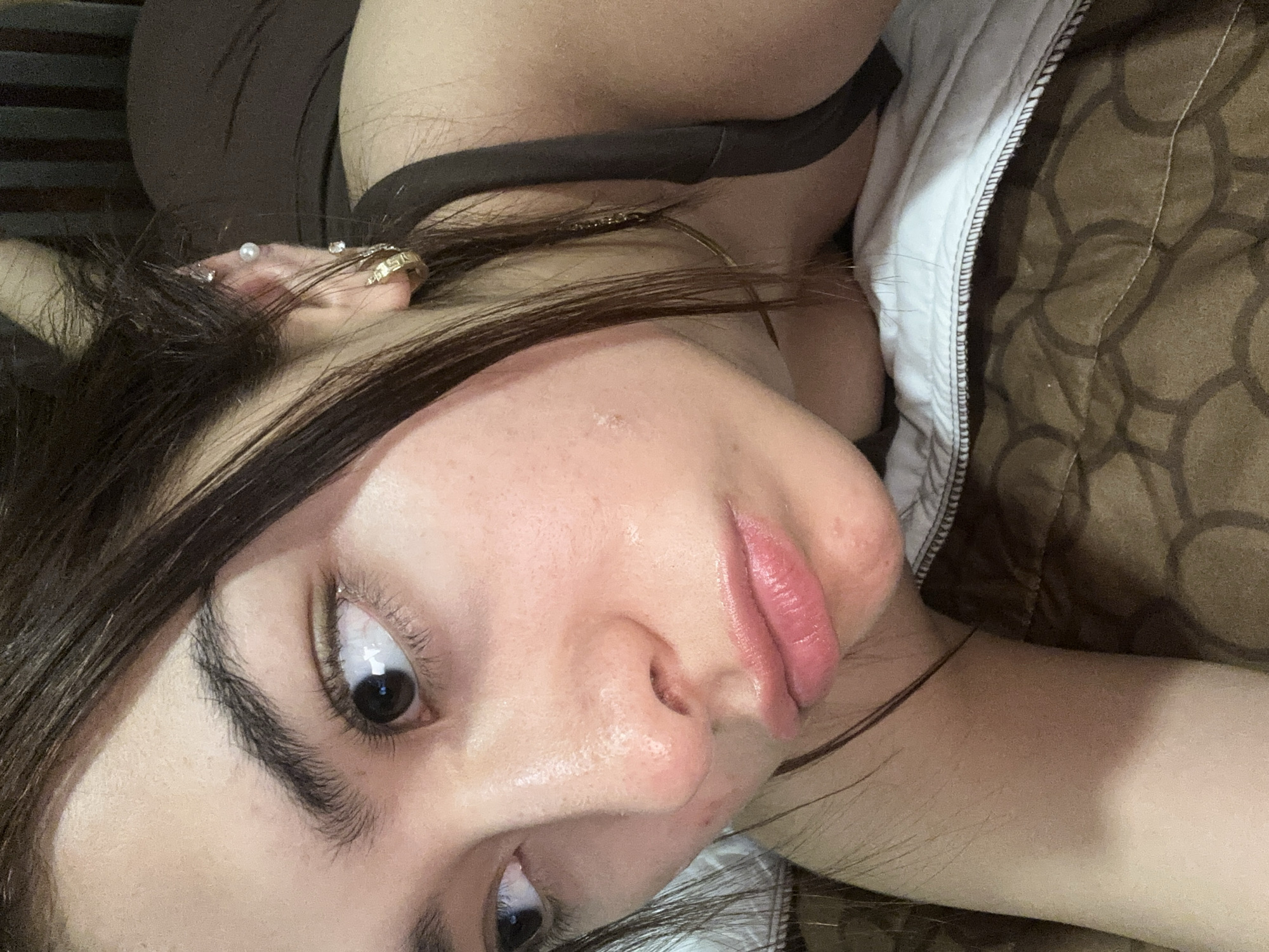 A close-up, angled view of a woman's face. She has dark hair, light skin, and prominent eyebrows. Her eyes are dark and directed upwards. She has multiple piercings in her left ear, including a pearl stud and a gold hoop earring. The skin on her face appears slightly dewy. She is lying on a bed with brown patterned bedding and a white edge visible near her neck. A dark-colored garment with a thin strap is also visible. The location is unknown; there is nothing in the image to indicate the city of Puebla, Mexico.