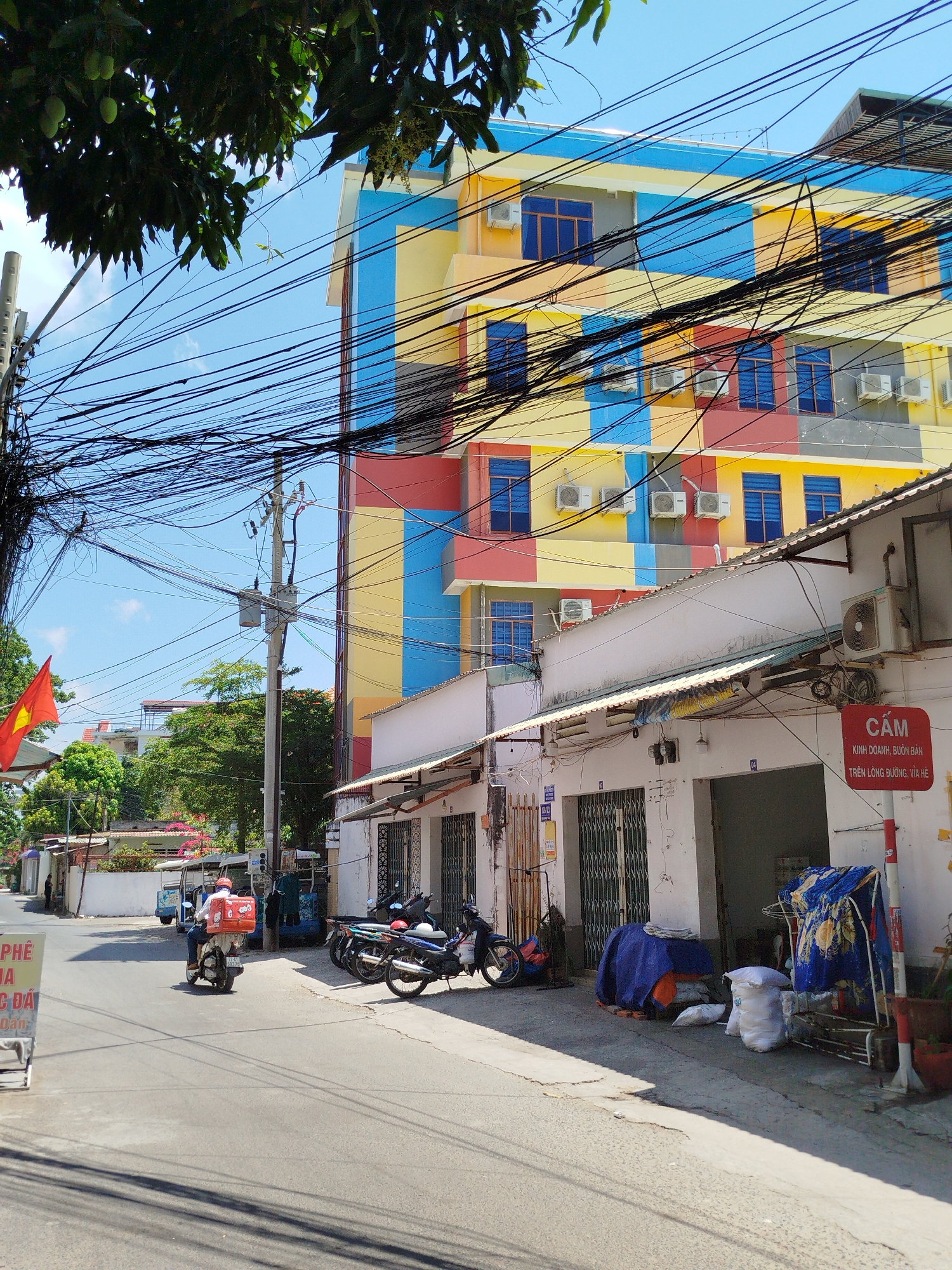 The scene is a street in Ho Chi Minh City, Vietnam, characterized by a mix of residential and commercial activity. A brightly colored building, painted in a patchwork of yellow, blue, red, and grey, dominates the background, with several air conditioning units attached to its façade. Tangled overhead power lines obscure parts of the sky and building.
Along the street, there are several one-story buildings with storefronts. Some are closed with metal gates, and the sidewalks are occupied by parked motorbikes, blue tarps covering various items, and sacks of goods. Signage, in Vietnamese, is visible on some of the buildings. The ground floor shops appear to be used for storage or small businesses. A sign warns against trading or business on the sidewalk.
A Vietnamese flag flies on the left side of the frame. A delivery driver on a scooter, identifiable by the orange delivery box on the back, is seen riding away from the viewer down the street. In the foreground, a sign advertising coffee is visible. The sky is clear and blue, indicating a sunny day.
