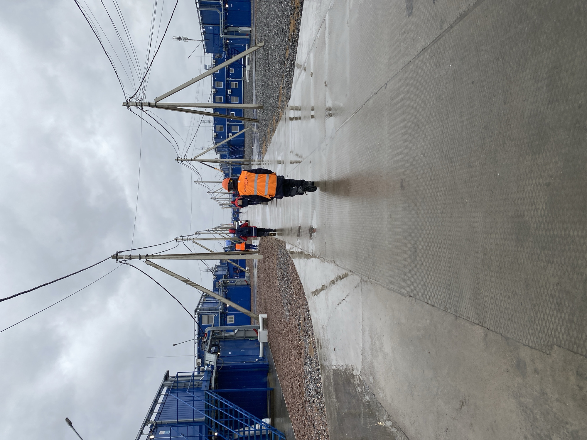 The image depicts a scene in Koskolovo, Russia, characterized by a row of blue, modular buildings lining a concrete walkway. The walkway is wet, reflecting the overcast sky above. Several people wearing orange safety vests are walking along the path. Overhead, utility poles with power lines are visible. Gravel is present between the buildings and the walkway. The overall atmosphere appears industrial.