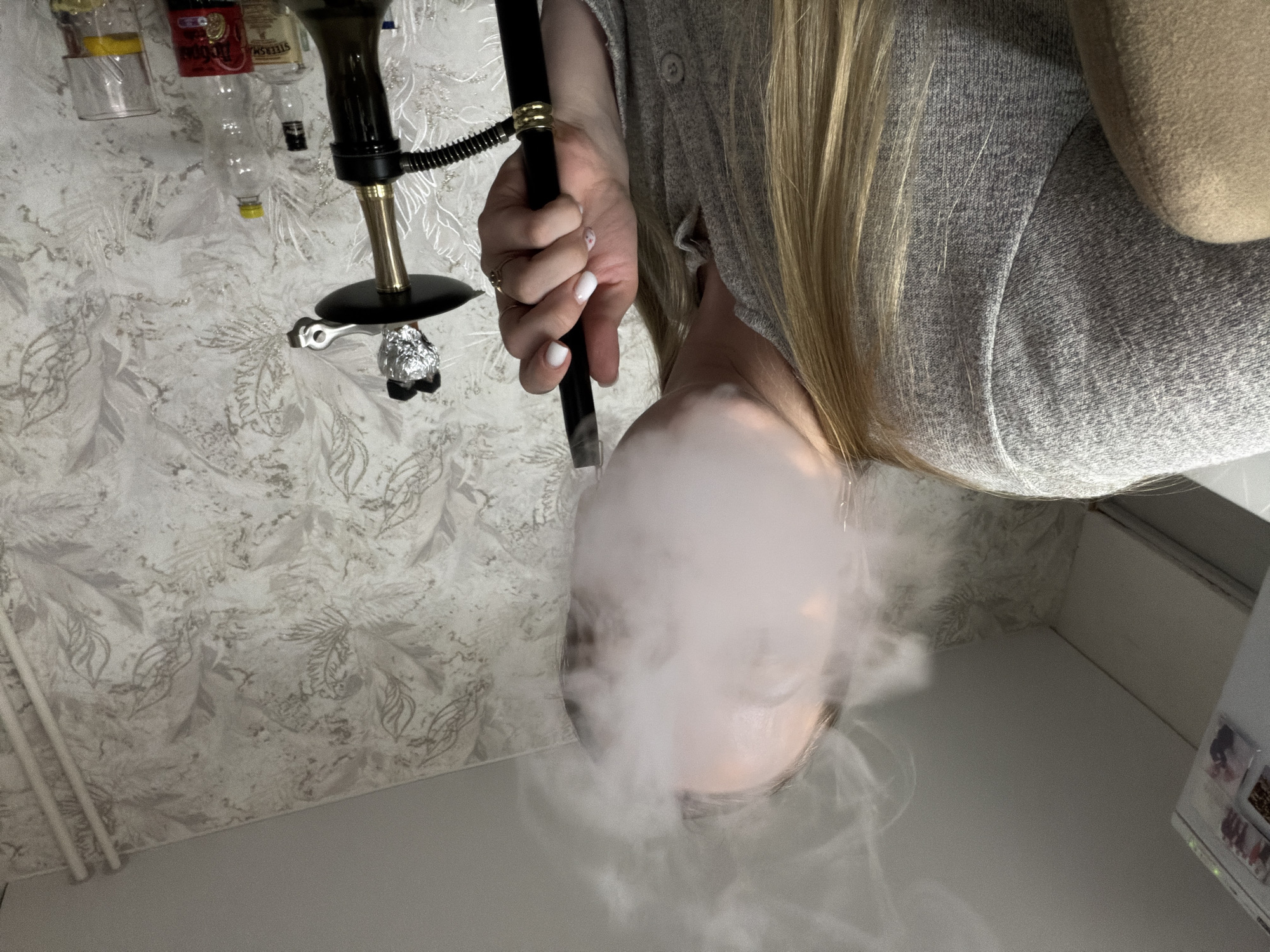 A woman with long blonde hair is exhaling smoke from a hookah. She wears a grey sweater and has white nail polish. The hookah has a black hose and gold accents. Behind her are several bottles, possibly alcoholic beverages, on a shelf. The background features a patterned wall. The location is unknown and cannot be determined from the image.
