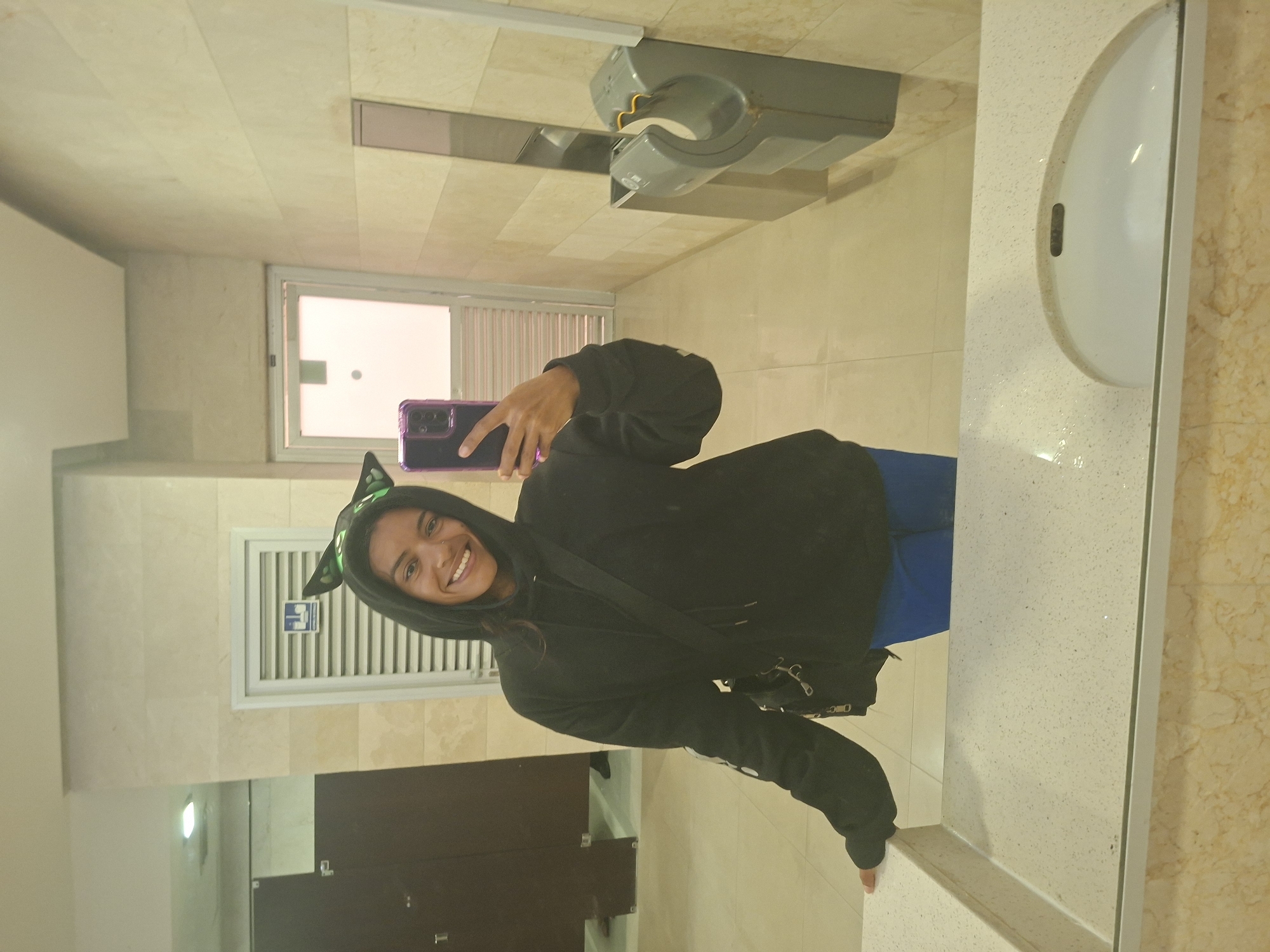 A person with dark hair and brown eyes, wearing a black hooded sweatshirt and blue pants, stands in a public restroom, taking a selfie in a mirror. They are wearing novelty cat ears and have a dark crossbody bag slung over their shoulder. The restroom has beige tiled walls and floors, with two dark-colored stalls visible in the background. A white sink with a speckled countertop is in the foreground, reflecting the person and the environment. A hand dryer is mounted on the wall to the right. The lighting appears to be overhead fluorescent lights, creating a bright and even illumination. The overall scene is typical of a public restroom, with no unusual or distinctive features that would tie it to a specific location beyond the city of Pampatar, Venezuela.
