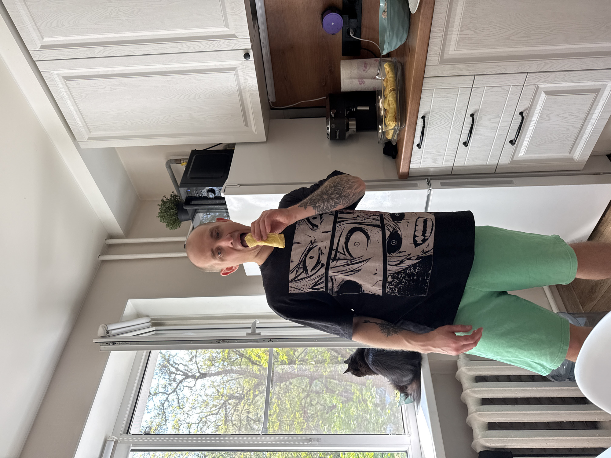 A man stands in a kitchen, holding a rolled food item in his mouth. He wears a black t-shirt with a manga illustration and light green shorts. His arms are tattooed. He stands near a window through which trees can be seen. A cat is perched on the window sill. The kitchen contains white cabinets with black handles, a white refrigerator, a microwave, and a coffee maker. There are rolled food items in a plastic container on the counter. The location is not specified.