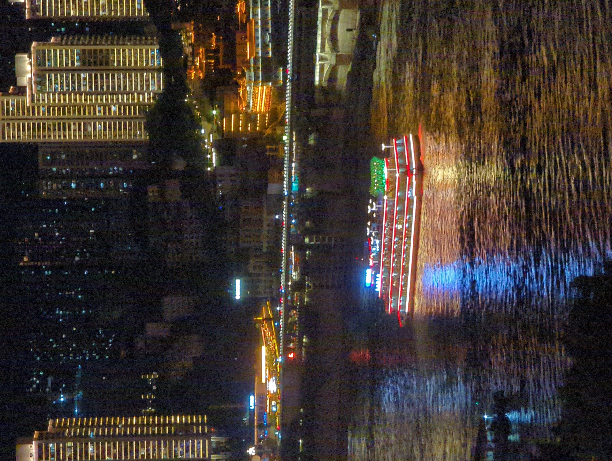 This is a nighttime, high-angle shot of a waterfront in Yuzhong District, China. The scene is dominated by the bright lights of a city skyline, featuring numerous illuminated high-rise buildings. In the foreground, a multi-tiered, illuminated riverboat, possibly a passenger cruise ship, is navigating a wide body of water. The boat has red and white lights outlining its decks and structure, and its lights reflect on the rippling water. In the middle ground, a bridge spans across the water, with lights from vehicles visible on its surface. Beyond the bridge, more city lights and buildings are visible, some with traditional architectural elements illuminated with warm light. The overall impression is a vibrant and active urban night scene.