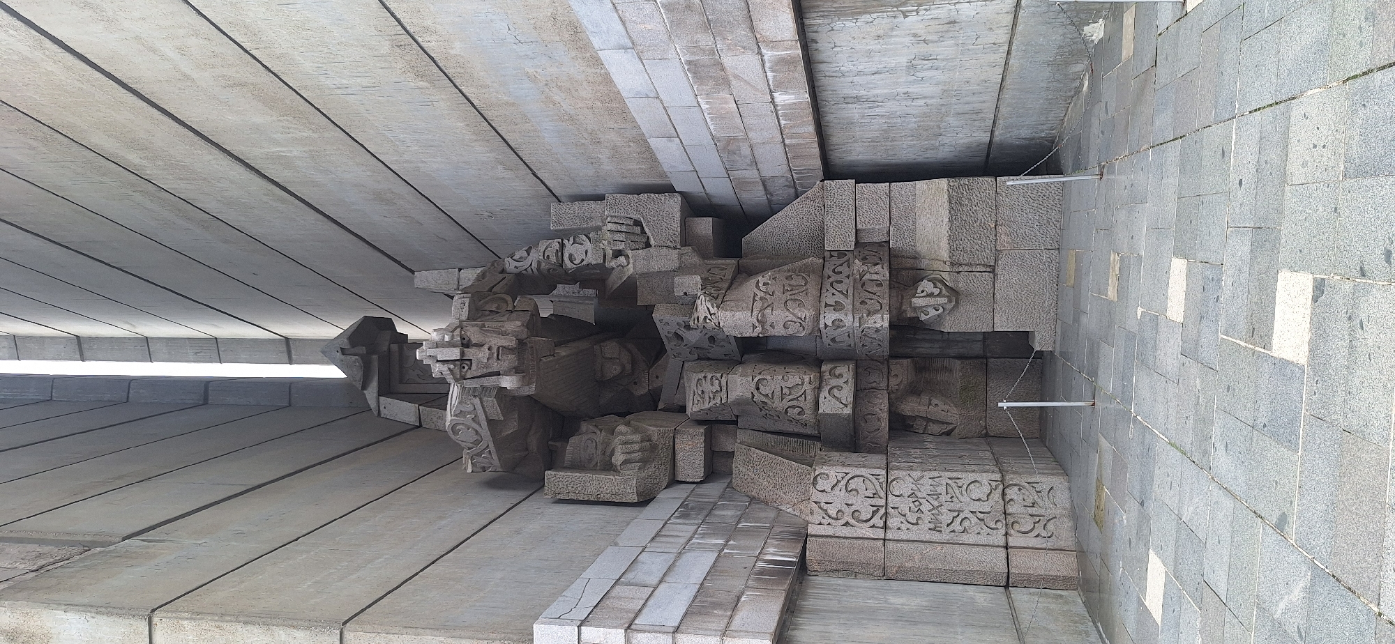 This image shows the Monument to the Founders of the Bulgarian State in Shumen, Bulgaria. The monument is carved into a hillside and features a large, abstract sculpture of a warrior. The sculpture is made of rough-hewn stone and is adorned with intricate carvings. The monument is surrounded by a paved plaza and a stone wall. The sky is visible in the background, and the sun is shining. The overall impression is one of grandeur and strength.