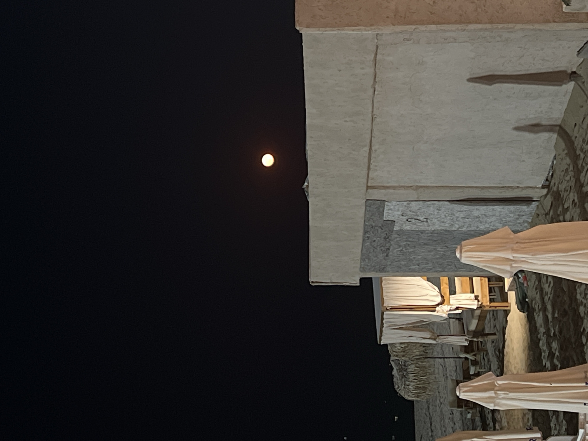 The image depicts a nighttime scene on a sandy beach, likely in Nesebar, Bulgaria. A bright, full moon is visible in the dark sky, casting a soft glow. In the foreground, several beach umbrellas are closed and covered with white fabric, appearing like shrouded figures. Beyond them, there are cabanas with white curtains, and wooden furniture. The structures in the background are made of concrete and have a rough, textured finish. The overall atmosphere is tranquil and serene.