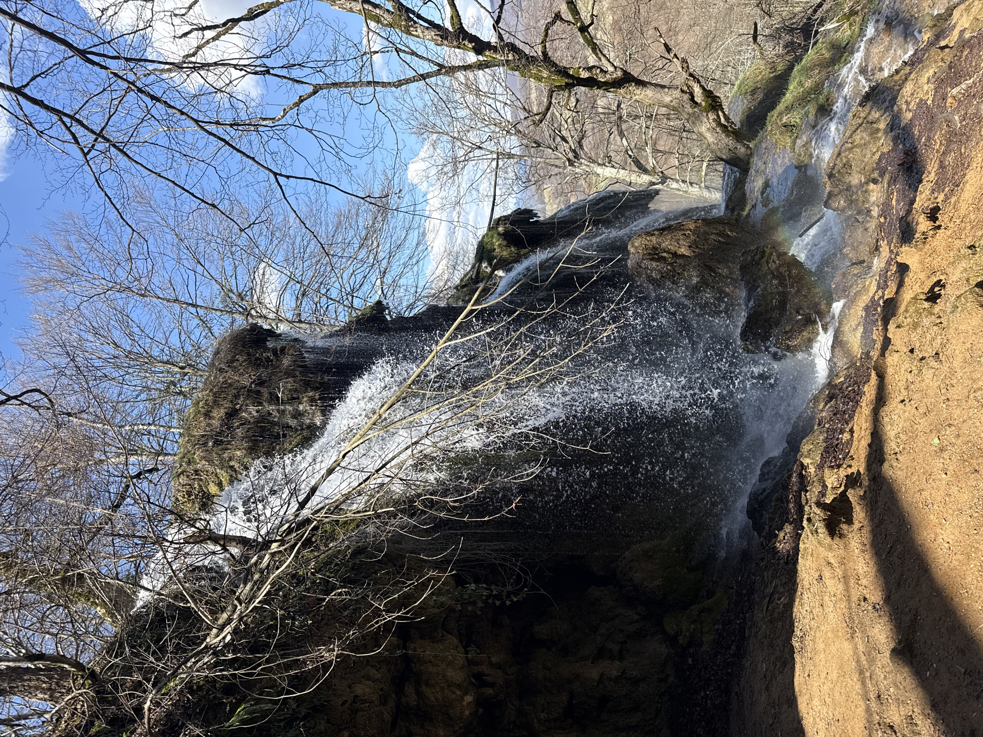 The image shows a waterfall, specifically the Skoka Waterfall in Ribaritsa, Bulgaria. Water cascades over a rocky cliff face, partially obscured by bare tree branches in the foreground. The sky is visible through the trees, with patches of blue and white clouds. The surrounding rock is brown and damp.
