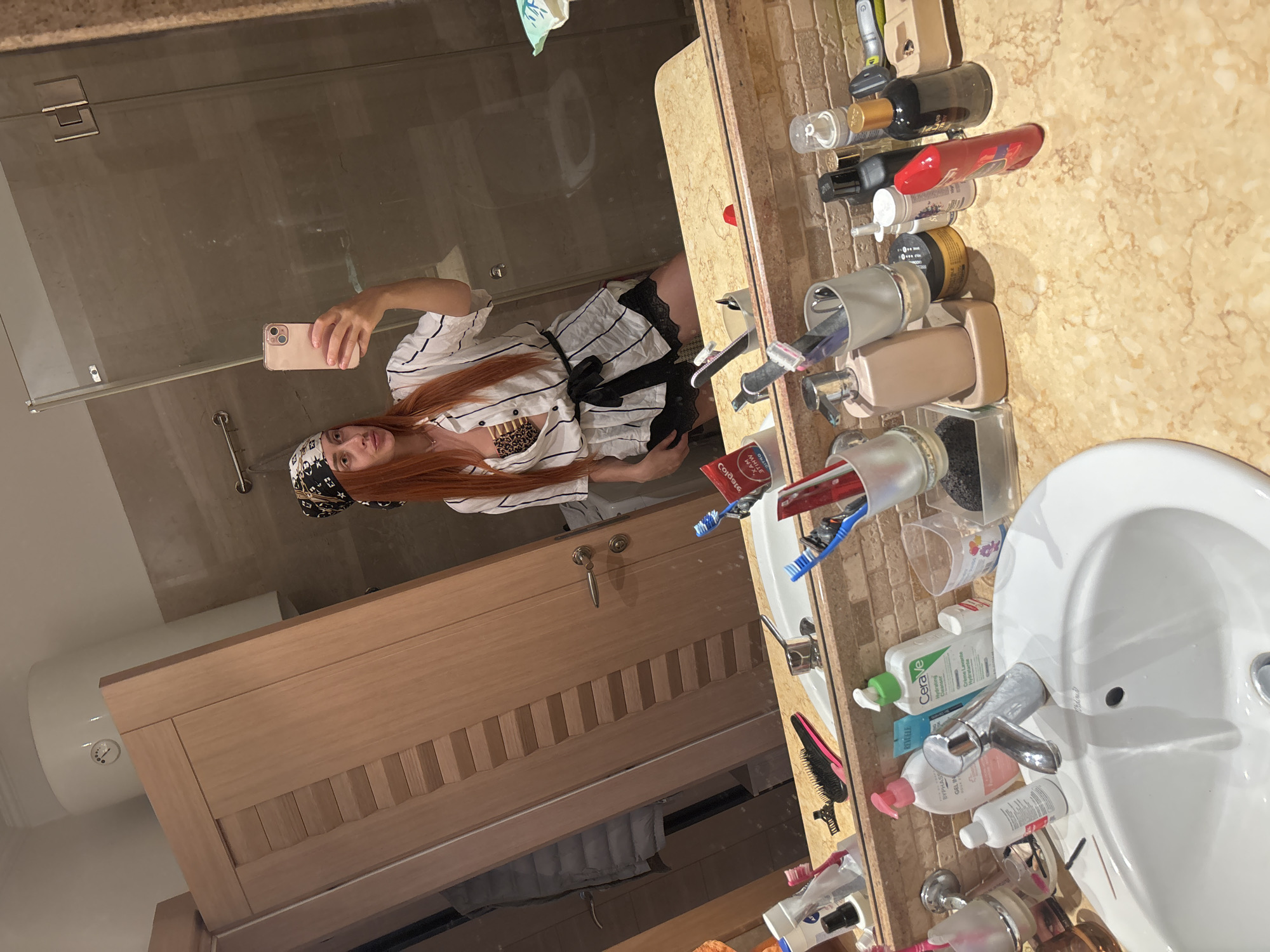 A woman with long red hair, wearing a black and white striped shirt, black shorts with lace trim, and a patterned bandana on her head, takes a mirror selfie in a bathroom. She is holding a pink smartphone in her right hand. The bathroom features a large mirror, wooden cabinets, and a vanity with a sink. The vanity is cluttered with various toiletries, including bottles of lotion, soap dispensers, toothbrushes, and a tube of toothpaste. The overall impression is that of a personal grooming space. The image context of Budva, Montenegro is not visually ascertainable from the image itself.