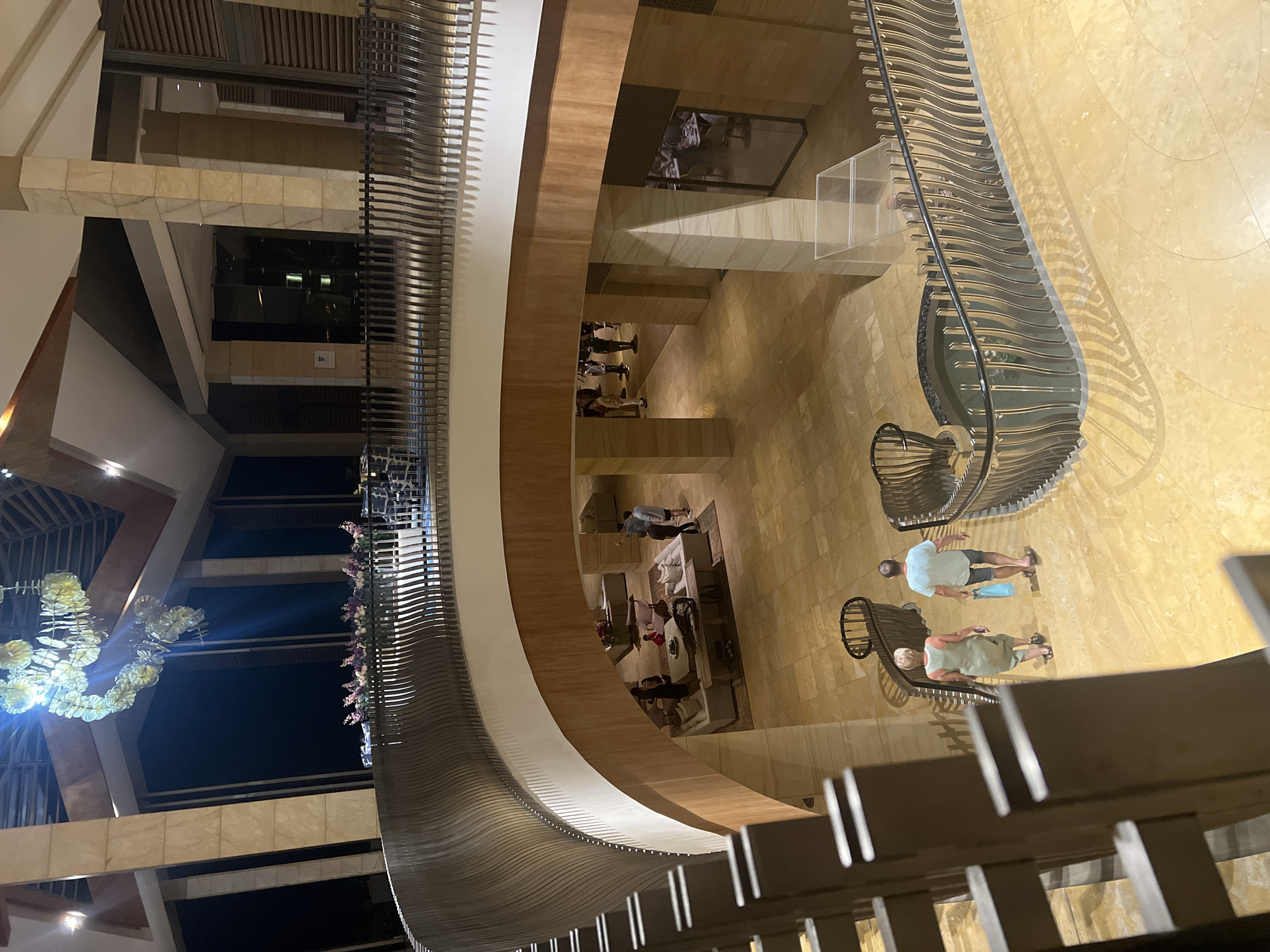 View of the interior of a multi-story building in Kuta Selatan, Indonesia. The interior is upscale, with marble floors and intricate metal railings. Several people are visible on different levels. A chandelier hangs from the ceiling. Tables and chairs are arranged on the lower level, suggesting a dining or seating area.
