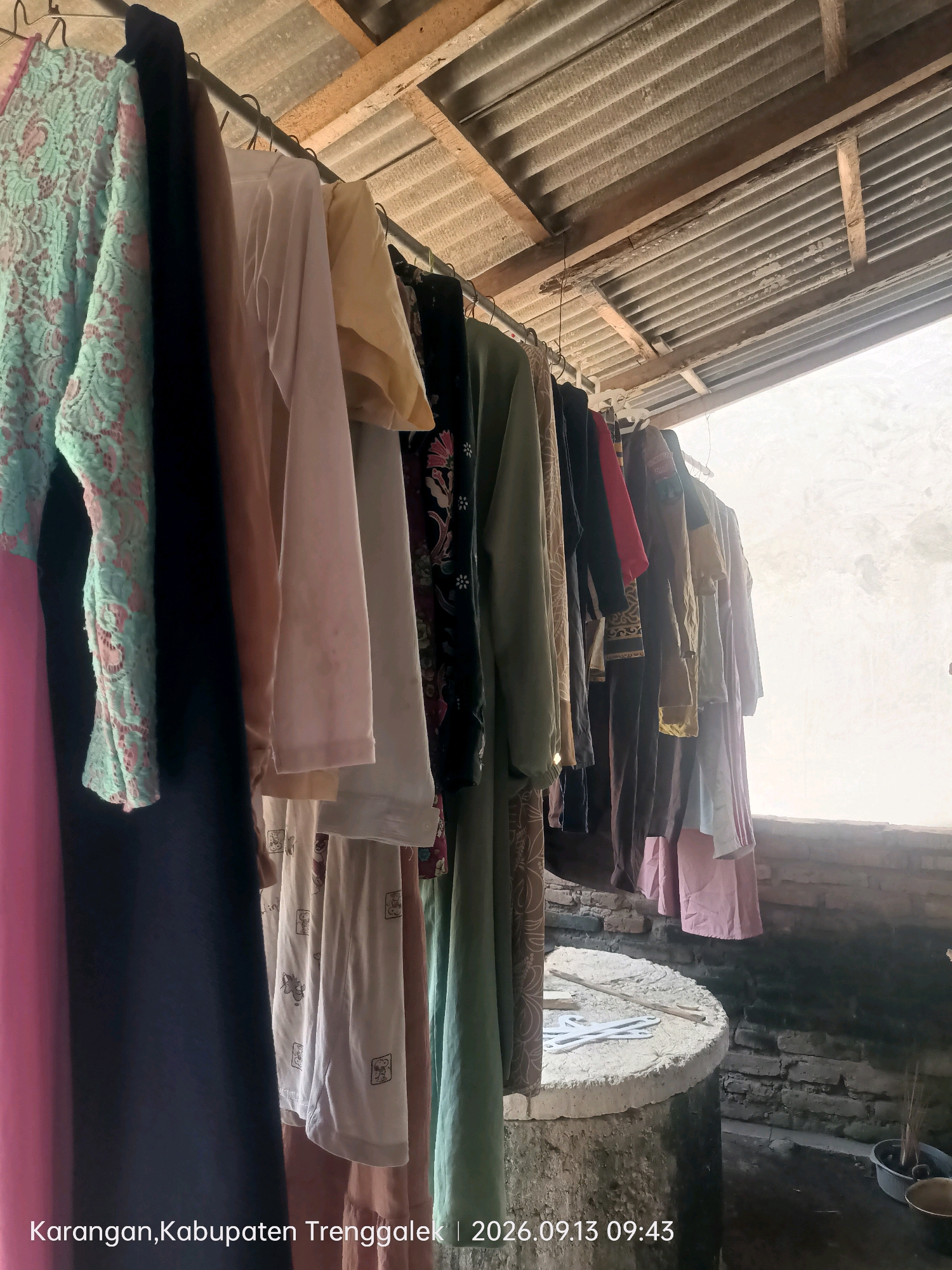 The image displays a close-up, eye-level view of various clothing items hanging on a clothesline. The garments are predominantly in muted colors such as beige, off-white, and various shades of brown and green. Some pieces feature subtle patterns, including what appears to be a floral or paisley print on one garment and a lace-like texture on another. The clothing is hung on metal hangers, which are suspended from a horizontal metal rod. Above the clothesline, a corrugated metal roof is visible, supported by wooden beams. The background is a bright, overcast sky, suggesting an outdoor setting. The bottom of the image shows a concrete well or cistern with a wooden stick resting on its rim. The surrounding area appears to be constructed of brick. Text overlays at the bottom of the image indicate the location as "Karangan, Kabupaten Trenggalek" and the date and time as "2026.09.13 09:43". This suggests the scene is in the Sumberingin area of Trenggalek Regency, Indonesia.
