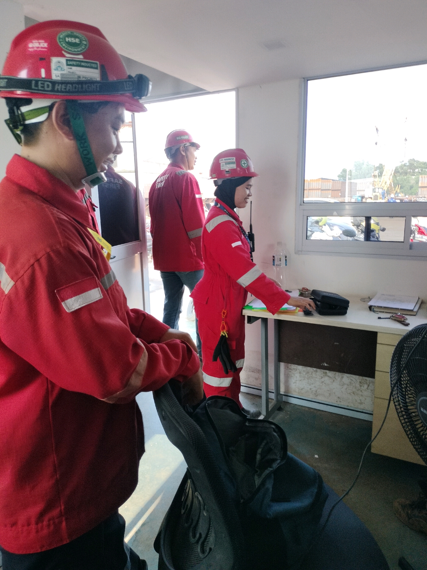 The image depicts an indoor scene with a bright, sunlit outdoor view visible through large windows. In the foreground, a person wearing a red hard hat with an attached LED headlamp and a red safety jacket is partially visible, seated in a dark chair. The jacket has reflective stripes and a small Indonesian flag patch on the left sleeve. Behind this individual, two other people in red safety outfits are present. One, a woman wearing a red headscarf and a red jumpsuit, stands at a white desk, interacting with a black device and papers. She has a radio clipped to her belt. Another person in a red jacket is standing further in the background. The environment appears to be a site office or control room, potentially at a construction or industrial facility in Batam, Indonesia, given the presence of construction equipment and cranes visible through the windows. The overall atmosphere suggests a work environment, possibly related to a project or operation in Batam.
