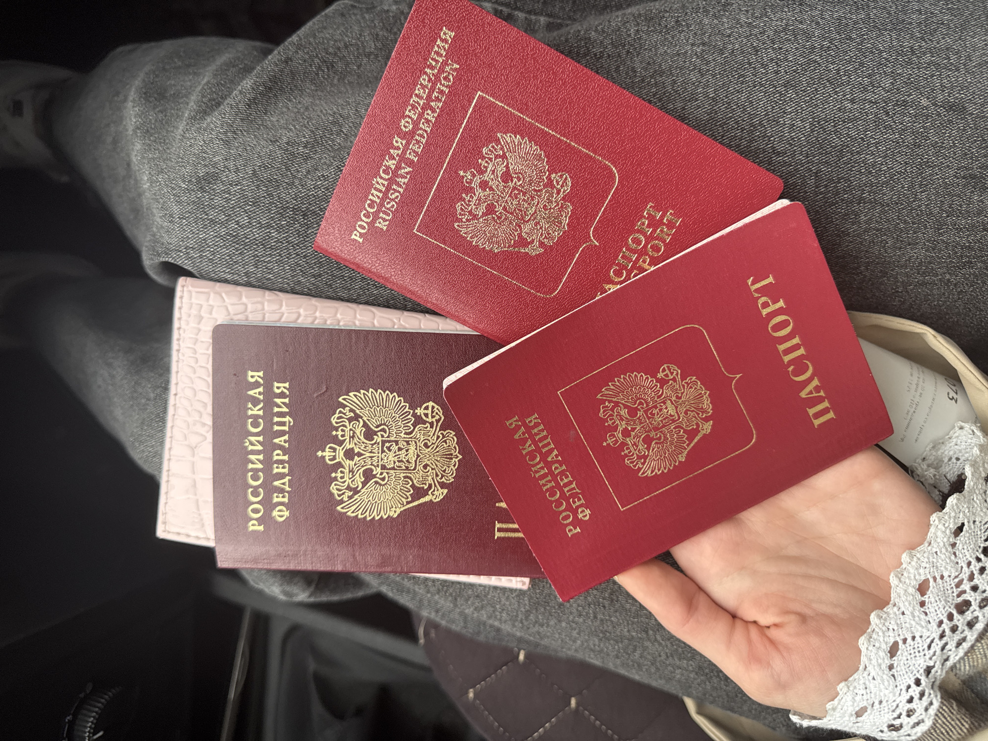 Three Russian Federation passports are fanned out on a person's lap. The passports are red and have the Russian coat of arms printed on them in gold. The person is wearing gray pants and a white lace cuff is visible on their wrist. The scene appears to be in the city of Perm, Russia.