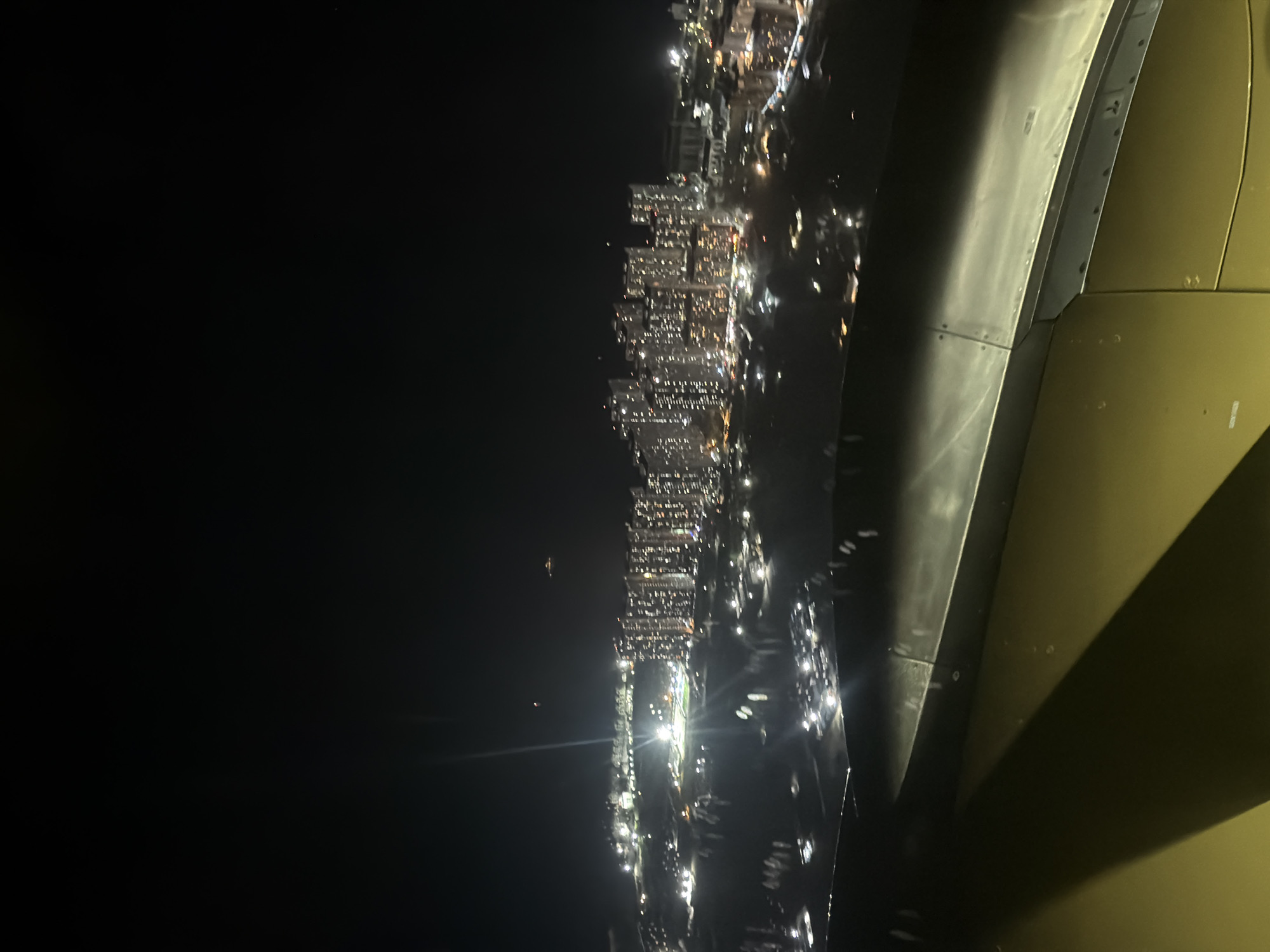 The image shows an aerial view of the city of Malé, Maldives, at night. The city is densely populated with brightly lit buildings. The scene is viewed from an airplane window, with part of the aircraft visible on the right side of the frame. The city lights illuminate the surrounding waters.