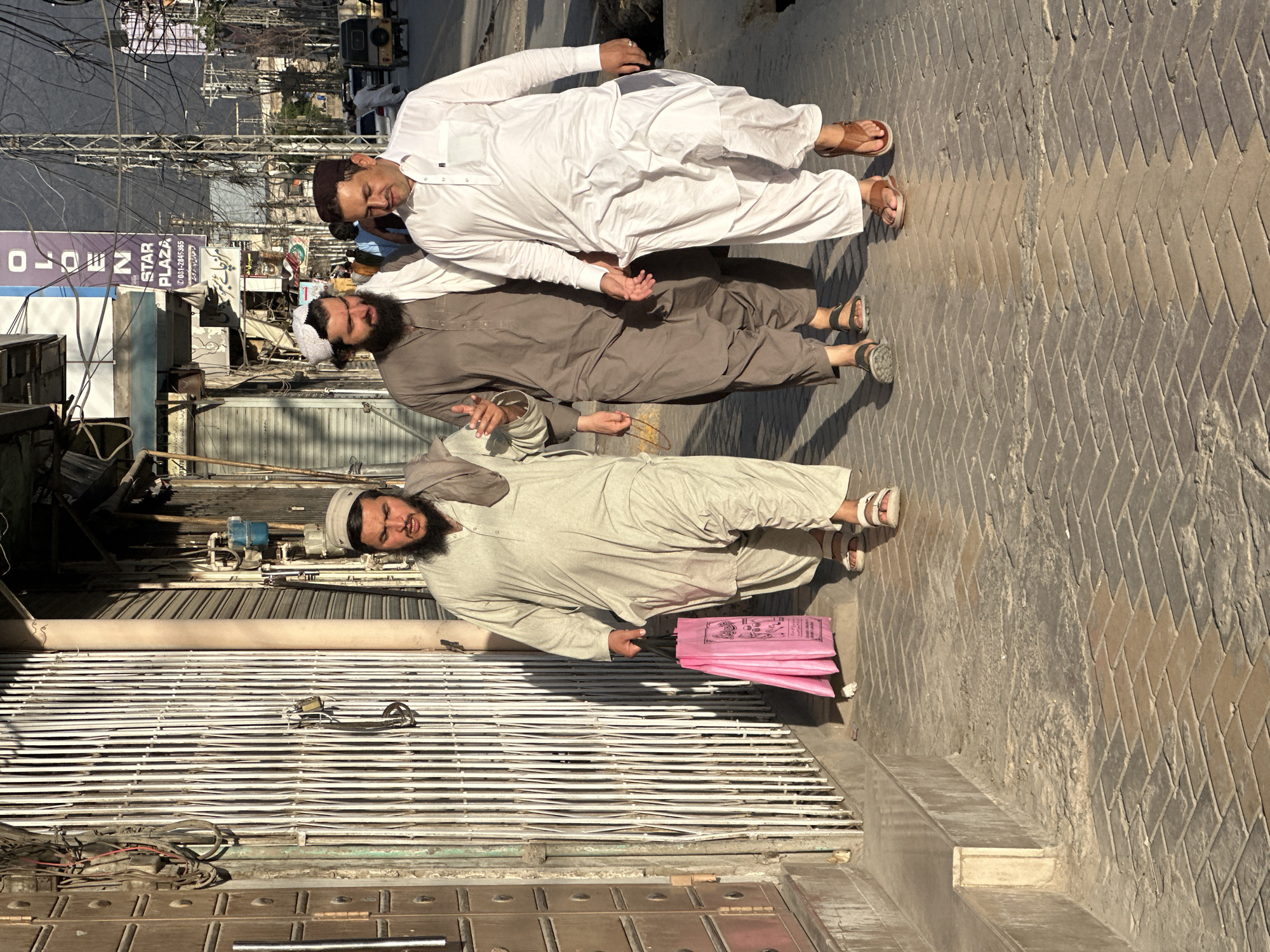 Three men stand in a row on a paved street. The man on the top is wearing a white shalwar kameez and a brown topi. The man in the middle is wearing a brown shalwar kameez and a white topi. The man at the bottom is wearing a light brown shalwar kameez and a white topi. He is also holding a stack of pink bags. They are standing in front of closed metal roll-up doors. Above the doors are electrical wires. A sign reading "Star Plaza" is in the background. The location is Quetta City Tehsil, Pakistan.
