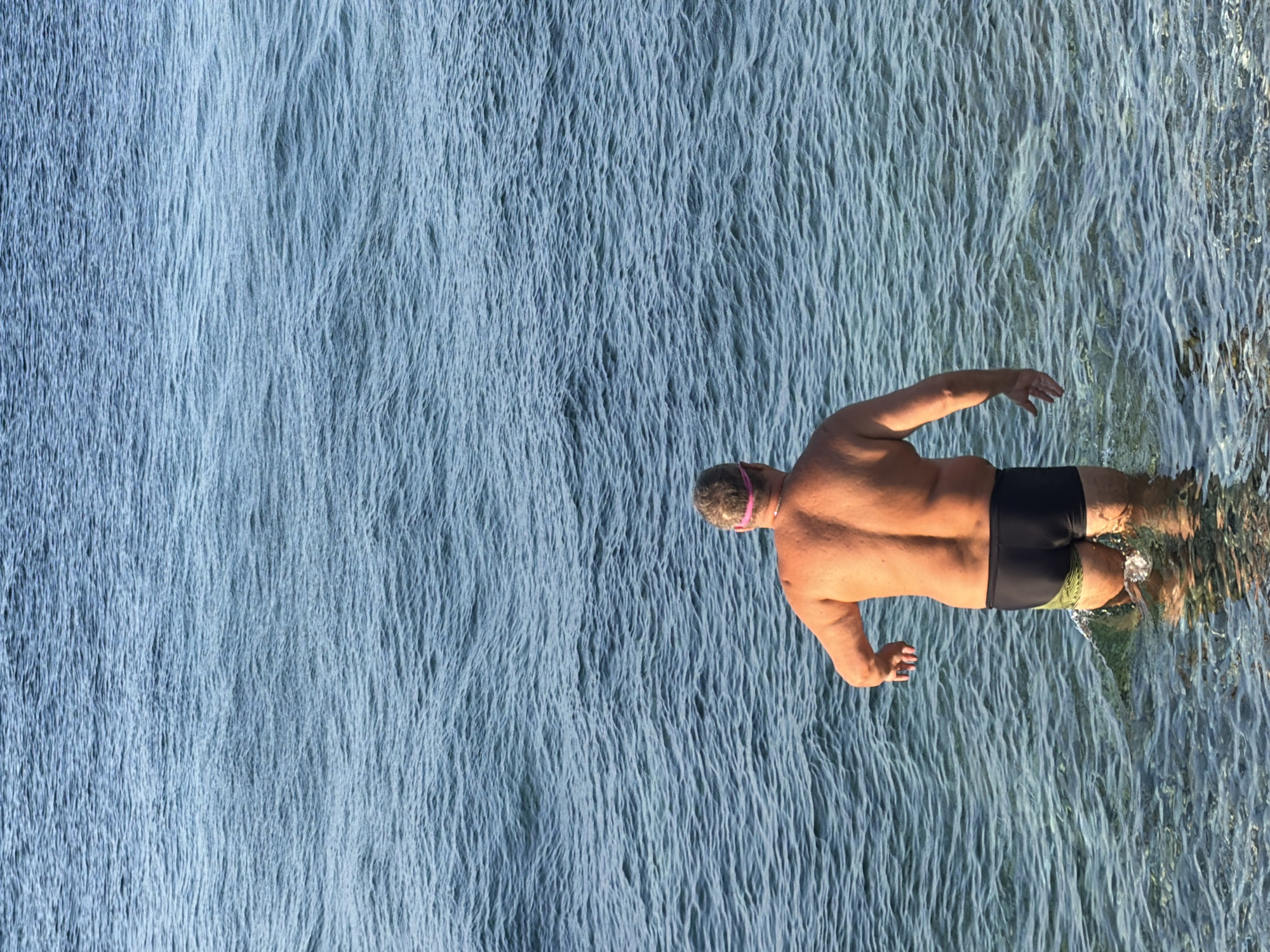 A middle-aged man with short, graying hair and a pink swimming cap is wading into the clear, blue water of the Adriatic Sea in Budva, Montenegro. He is wearing black swimming trunks with a green and yellow striped accent on the sides. His back is to the camera, and he is facing towards the open sea. The water is calm, with small ripples on the surface, and the seabed is visible beneath the shallow water. The lighting suggests it is daytime, possibly late afternoon or early morning due to the warm glow on the man's skin.
