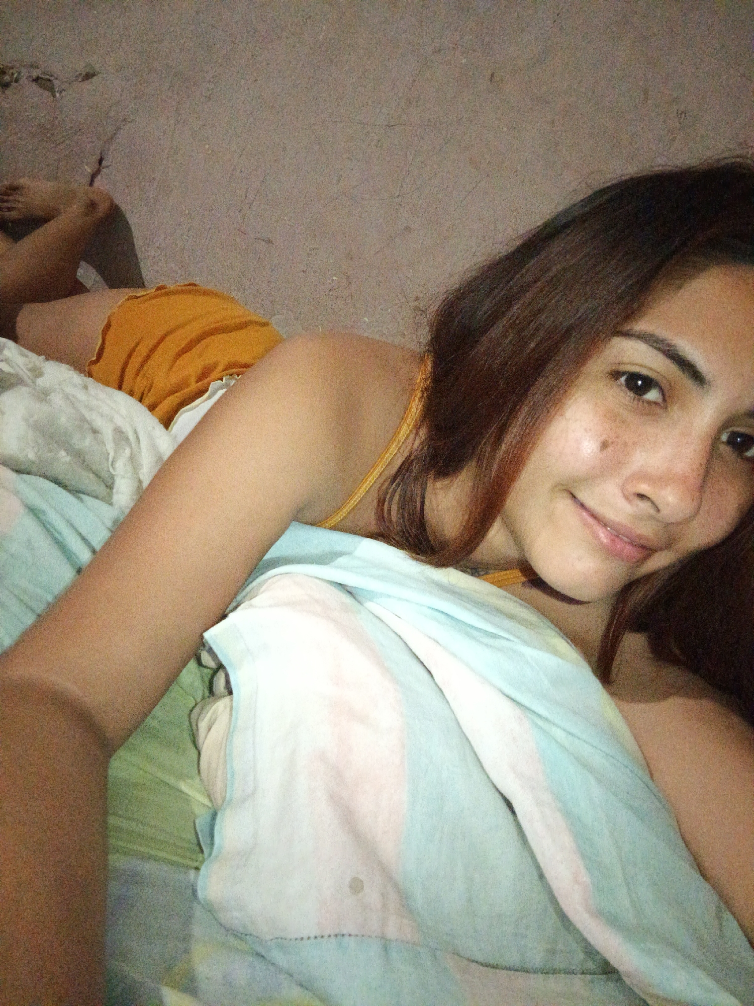 A young woman with brown hair and freckles lies on a bed in a room. She is wearing orange shorts and a yellow top. The walls of the room are made of concrete. The bed is covered with light blue, white, and pink striped bedding. The scene appears to be taken from a low angle, with the woman looking directly at the camera and smiling. The lighting is dim, suggesting it may be nighttime or a dimly lit room. The location is identified as Juan Griego, Venezuela.