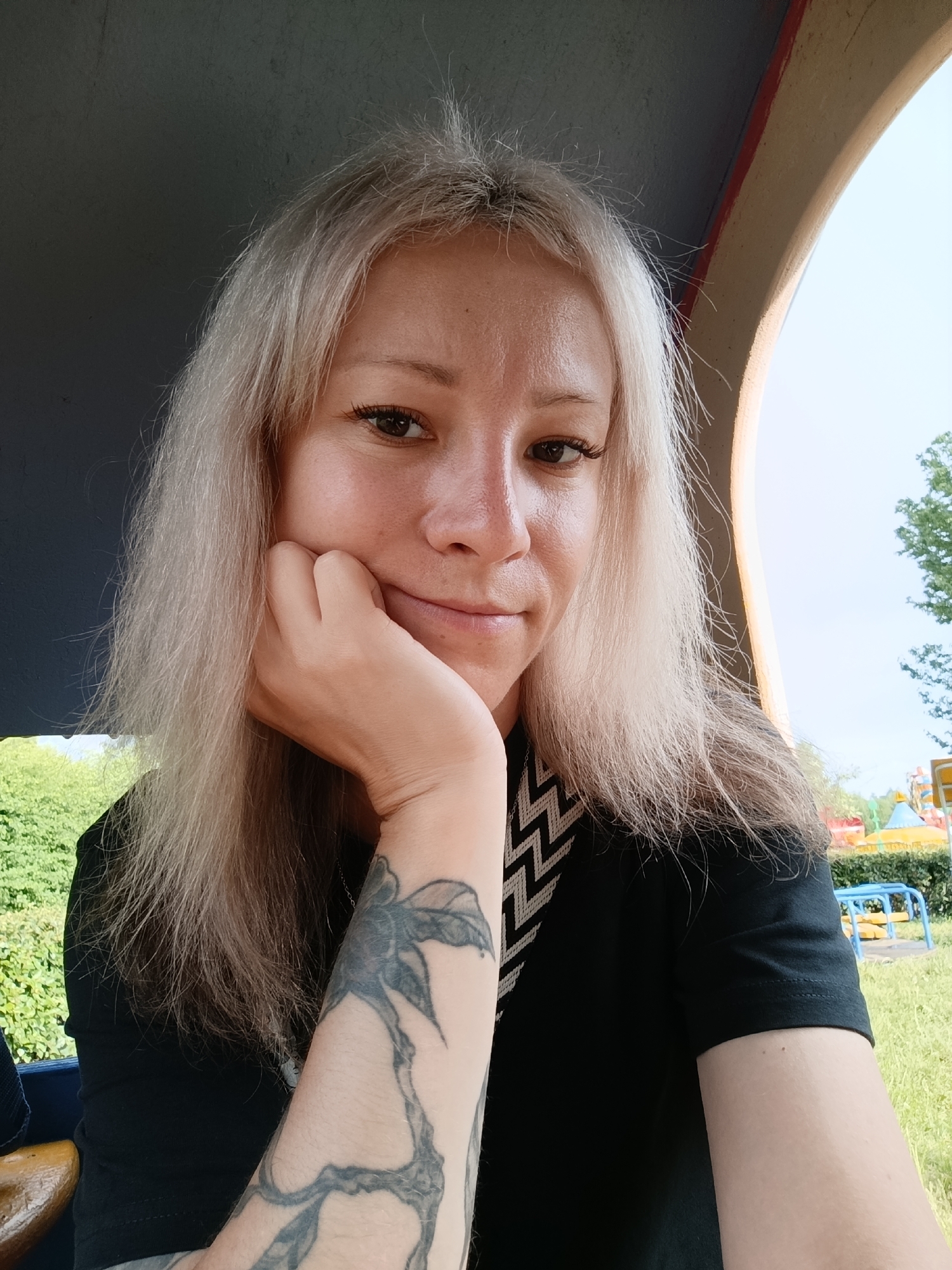 The media file is a selfie of a woman with blonde hair, wearing a black t-shirt. She is resting her chin on her hand, and her arm, which has a large tattoo of what appears to be a floral design, is visible. The background suggests an outdoor setting, possibly a park or playground, with green foliage and a bright sky. There are some colorful structures visible in the distance, and a hint of a playground set. The lighting suggests it is daytime, likely sunny. The woman has a relaxed and contemplative expression. No visible text is present in the image.