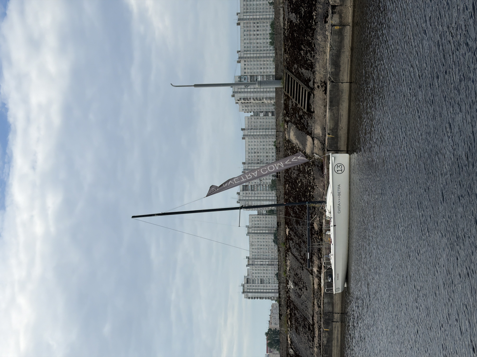 This is a full-frame, eye-level, outdoor, medium shot of a white sailboat docked in a canal with Saint Petersburg's residential buildings in the background. The sailboat is white with a black mast and a dark gray sail that has white text on it, which reads "СИЛА >>> ВЕТРА" and has a circled number 13. The building in the background are grey, multi-story apartment buildings. The sky is cloudy and blue with some white clouds. The water in the canal is dark and choppy. A concrete embankment with some sparse vegetation is visible along the edge of the canal. Wooden stairs lead up the embankment from the canal. The location is a canal in Saint Petersburg, Russia, likely near one of the city's residential districts. There are no people visible in the image.