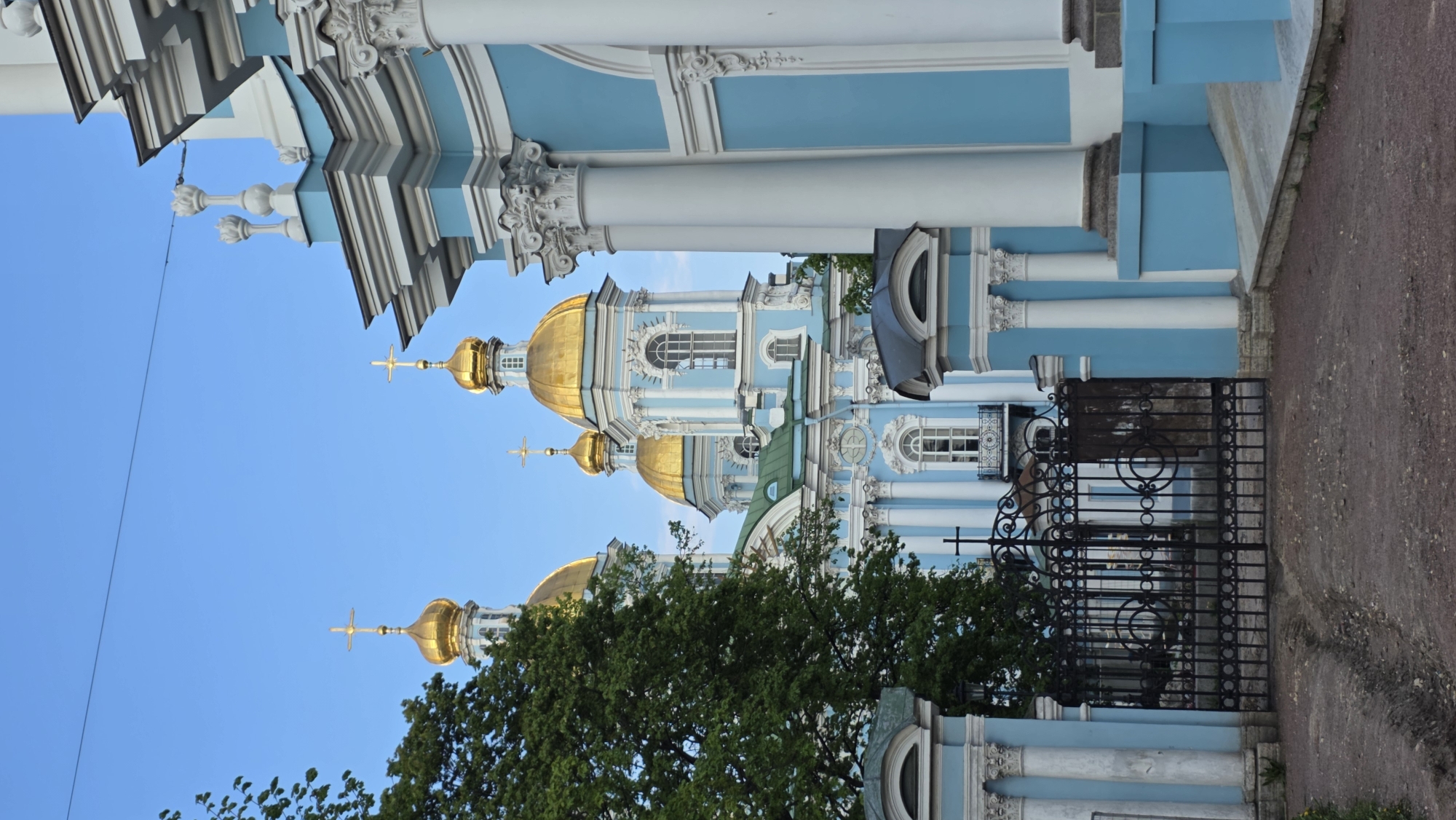 The image shows a low-angle view of the Smolny Cathedral in Saint Petersburg, Russia. The architectural style is Baroque, characterized by its light blue and white color scheme, ornate decorations, and golden domes topped with Orthodox crosses. Three golden domes are partially visible, with the top dome reaching into a clear blue sky. The cathedral's facade is detailed with columns, decorative moldings, and window frames. In the foreground, a black wrought-iron fence runs alongside a pavement. A tree with dense green foliage partially obscures the lower part of the building. The image is slightly tilted.