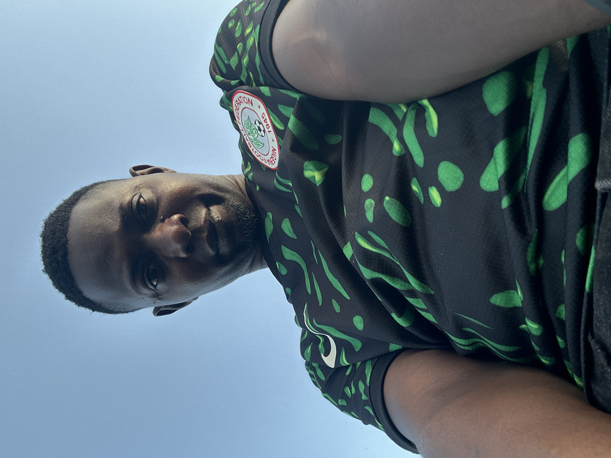 A low-angle, waist-up, outdoor selfie is captured against a clear blue sky. The subject is a Black man wearing a black and green patterned Nigeria national football team jersey. He has short, dark hair and a light beard. His left arm and shoulder are visible in the foreground, and he is looking down at the camera. The image is sharp and well-lit, with no apparent distortions.