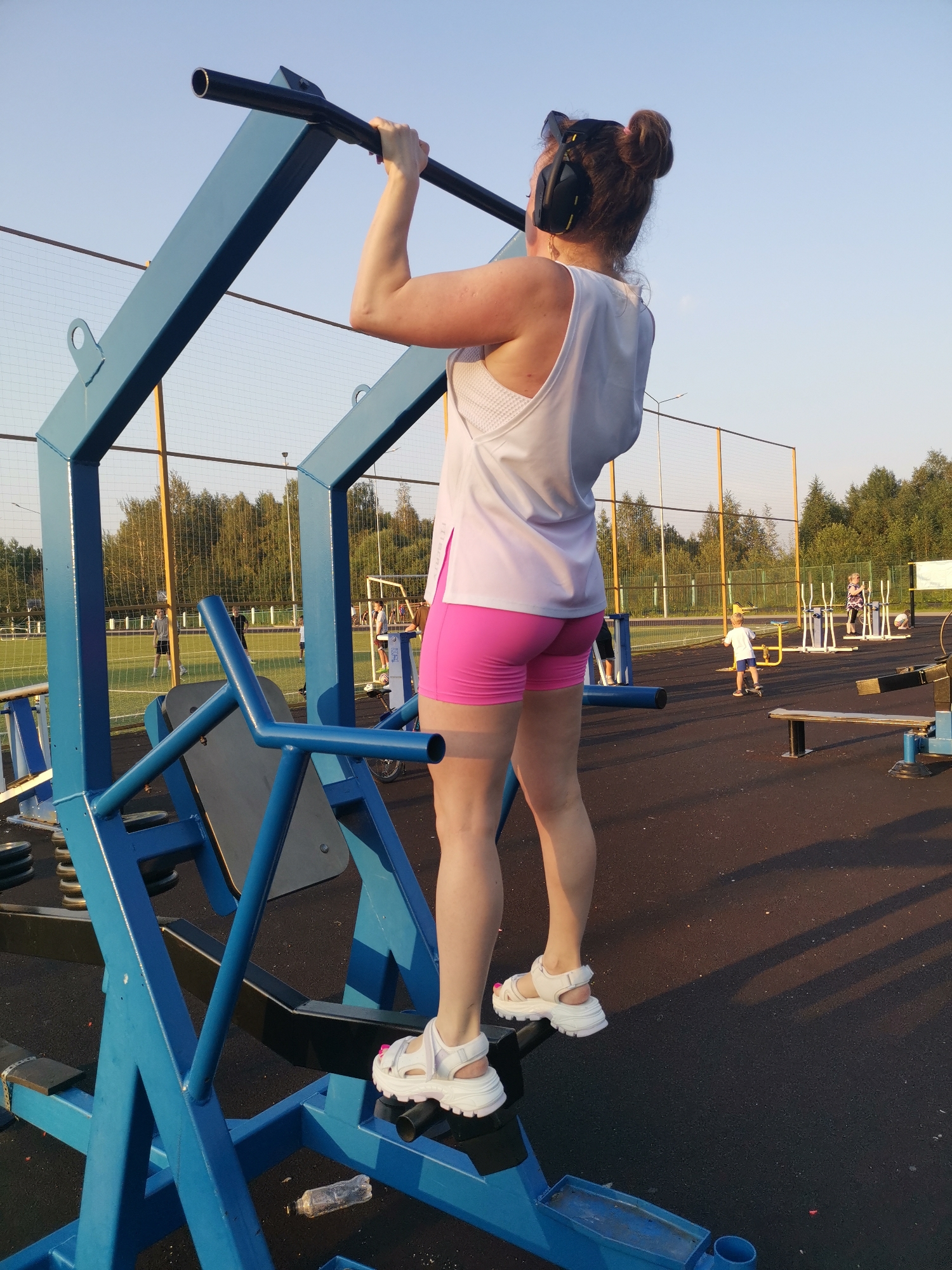 In Kizel, Russia, on a sunny day, a woman stands on an outdoor exercise machine, gripping a horizontal bar above her head. She is wearing a white tank top, pink shorts, and white sandals. Her hair is styled in two buns. She has headphones on and sunglasses on top of her head. The exercise machine is blue and made of metal. In the background, other people are exercising on similar equipment, and a soccer field is visible. The ground is covered with a dark, rubberized surface. A discarded plastic bottle lies on the ground near the exercise machine.