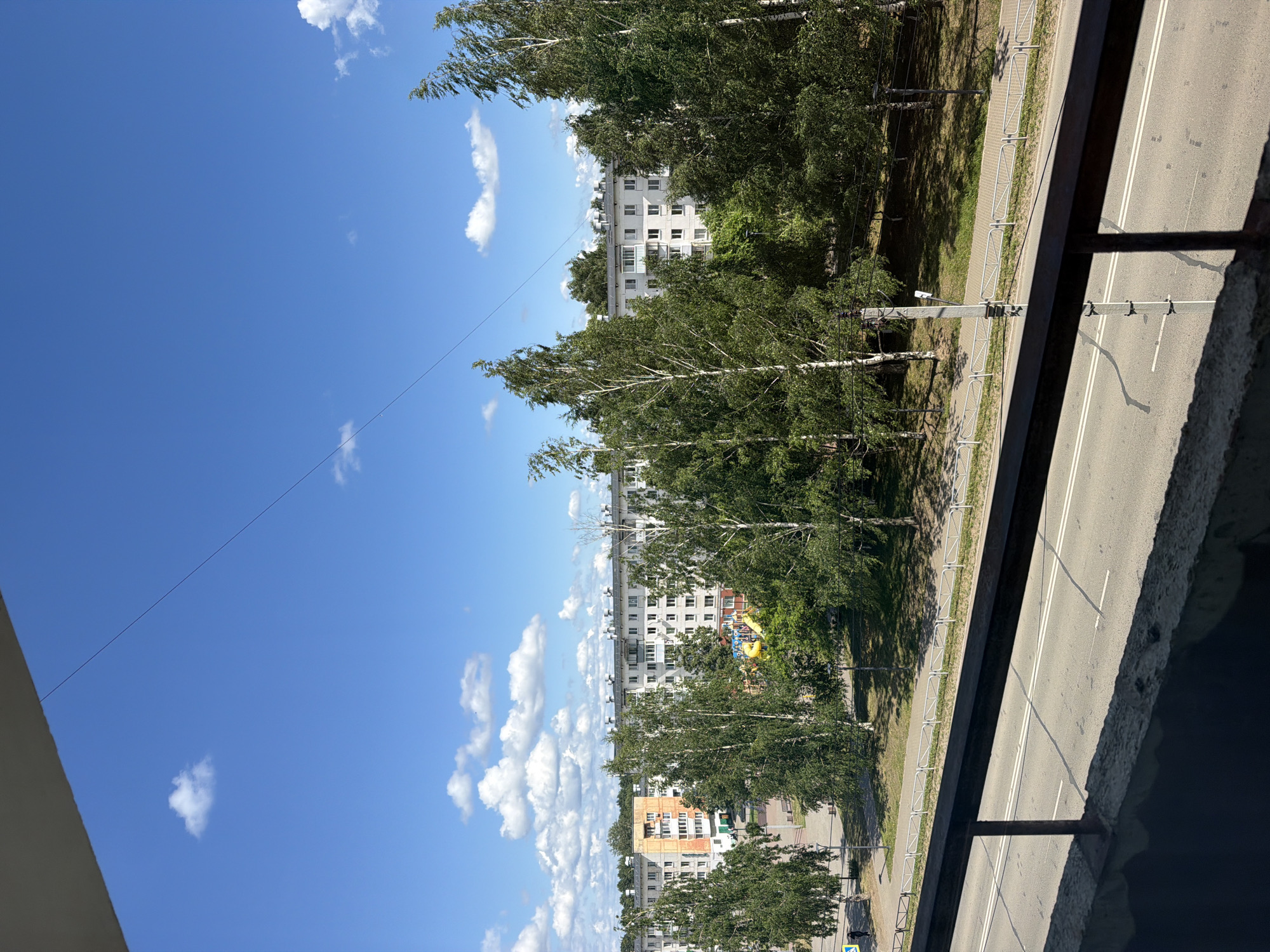 The image displays a street scene in Chaikovsky, Russia, on a bright, sunny day. The perspective is from a balcony or elevated window, looking down and across at a road lined with trees and apartment buildings. The sky is a vibrant blue with scattered white clouds, indicating pleasant weather.
In the foreground, a concrete balcony railing casts shadows onto the road. The road itself is asphalt with clear white lane markings. Adjacent to the road is a sidewalk, separated by a low, silver metal fence. Beyond the sidewalk, a strip of green grass is shaded by a dense line of mature trees, primarily birch trees with their distinctive white bark and lush green foliage.
Behind the trees, several multi-story apartment buildings are visible. These buildings appear to be of Soviet-era architecture, with numerous windows and balconies. One building has some colorful playground equipment visible in front of it, suggesting a residential area. The overall setting is urban with a significant amount of greenery.
There is no discernible activity or event taking place in the image, such as people or vehicles in motion. The scene appears to be a still, everyday view of the neighborhood. The time of day is likely midday or early afternoon, given the bright sunlight and the sharp shadows. No text is visible in the image.