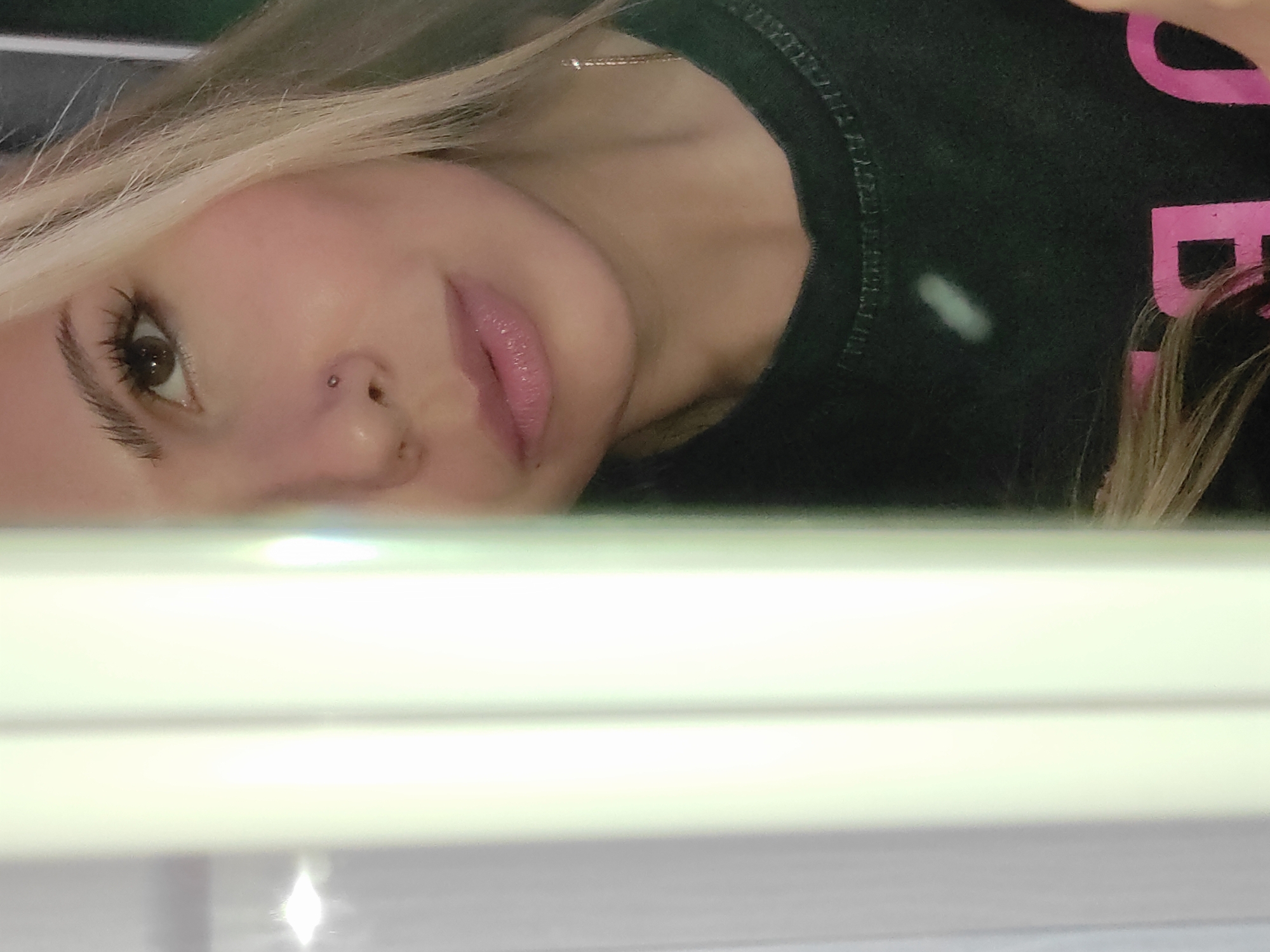 The image presents a close-up, rotated view of a person's face and upper body, seen from a slightly upward angle, as if looking into a reflective surface. The subject is a person, likely female, with long, blonde hair that partially covers the dark background. Her right eye is prominent, featuring a dark iris, long dark eyelashes, and a well-defined eyebrow. A small, silver-colored piercing is visible on her right nostril. Her lips are slightly parted and appear to have pink lip gloss.
She is wearing a dark-colored t-shirt, possibly black or dark green. On the t-shirt, near her left shoulder, a faint, light-colored mark or stain is visible. Partial pink text is also discernible on the t-shirt, showing the letter "D" and a portion of another letter. A delicate, gold-colored chain necklace is visible around her neck. The lower half of the image is dominated by a bright, white, and reflective surface, likely a mirror or a polished table, displaying several bright light reflections. The overall impression is that the person is taking a selfie or simply observing her reflection. Her expression is neutral and calm. The setting appears to be indoors, with no specific outdoor or geographical cues, though the location context provided is Olkhovka, Russia.