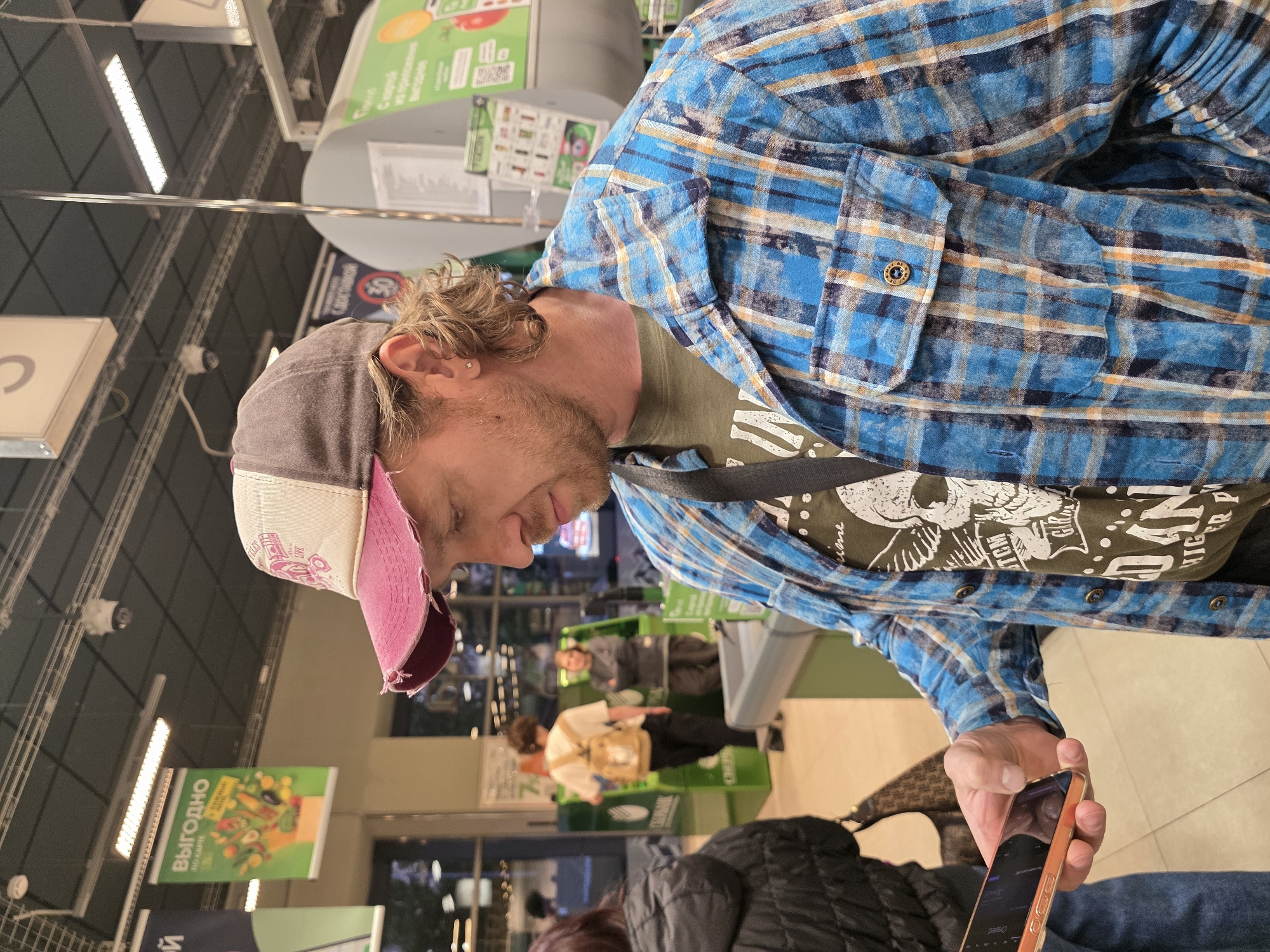 This image depicts an indoor scene, likely a supermarket or grocery store in Saint Petersburg, Russia. The primary subject is a man, possibly in his 20s or 30s, with light brown, slightly messy hair and a beard. He wears a gray baseball cap with a worn pink brim, a blue, orange, and black plaid long-sleeved shirt unbuttoned over an olive green t-shirt with a graphic design (partially showing a skull, "TCM", "DAN", and "HIGHER P"). He also has a small earring in his left ear.
The man is seen from the side, looking down intently at a gold-colored smartphone held in his right hand. His expression appears focused and neutral. In the background, other customers and green self-checkout machines are visible, suggesting a typical retail environment. The ceiling features bright, linear fluorescent lights and what appear to be security cameras.
Prominent Cyrillic text on various hanging signs confirms the Russian location. These include "ВЫГОДНО ПО КАРТЕ" (ADVANTAGEOUS WITH CARD), "С ДОСТАВКОЙ" (WITH DELIVERY), and "С картой приложения Выгоднее" (With the app card, it's more advantageous/profitable). The logo for "СБЕРБАНК" (SBERBANK), a major Russian bank, is also visible on one of the signs. A button on the man's sleeve reads "Created". The image captures a routine moment in a bustling public space, with the man engaged with his mobile device.