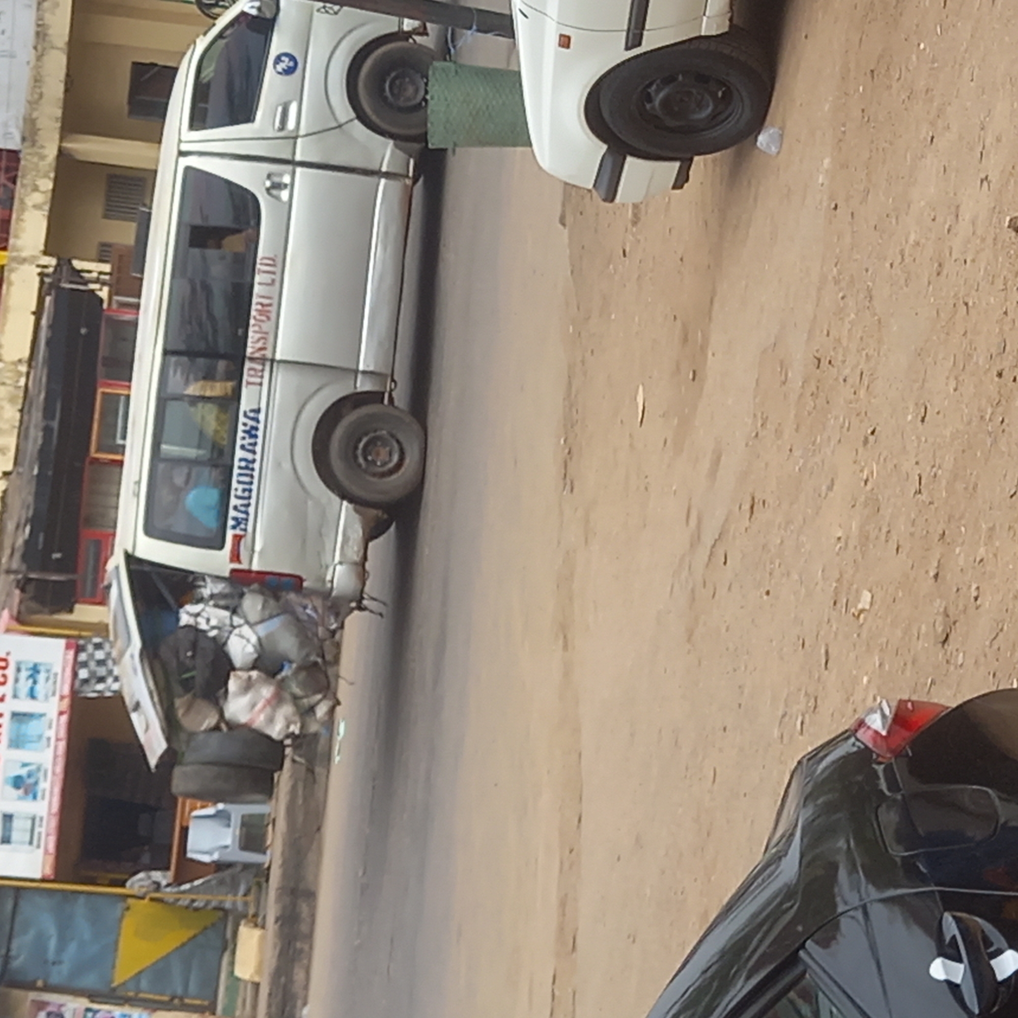 The image depicts a street in Dadi Riba, Nigeria. A white van labeled "MAGORAWA TRANSPORT LTD" is parked on the side of the road. The back of the van is open and filled with what appears to be various objects wrapped in cloth. A portion of another vehicle is visible in the bottom right corner of the frame. A few buildings are visible in the background, including one with a store sign. The ground appears to be a mix of paved road and dirt.