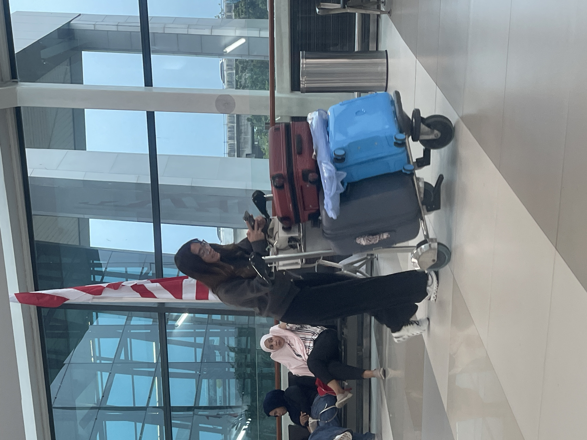 The image depicts a woman with dark hair, wearing glasses, a grey long-sleeved top, and black pants, walking through an airport terminal. She is pushing a luggage cart laden with three suitcases: a red one on top, a dark grey one below it, and a bright blue one at the bottom. In the background, two women are seated on a bench. One is wearing a dark hijab and the other a light pink hijab and a striped shirt. A Canadian flag is visible behind the walking woman, near a large window that offers a view of the sky and an elevated structure. The floor is tiled in a light and dark grey pattern. This scene is consistent with the interior of Soekarno-Hatta International Airport, located in Tangerang, Indonesia.