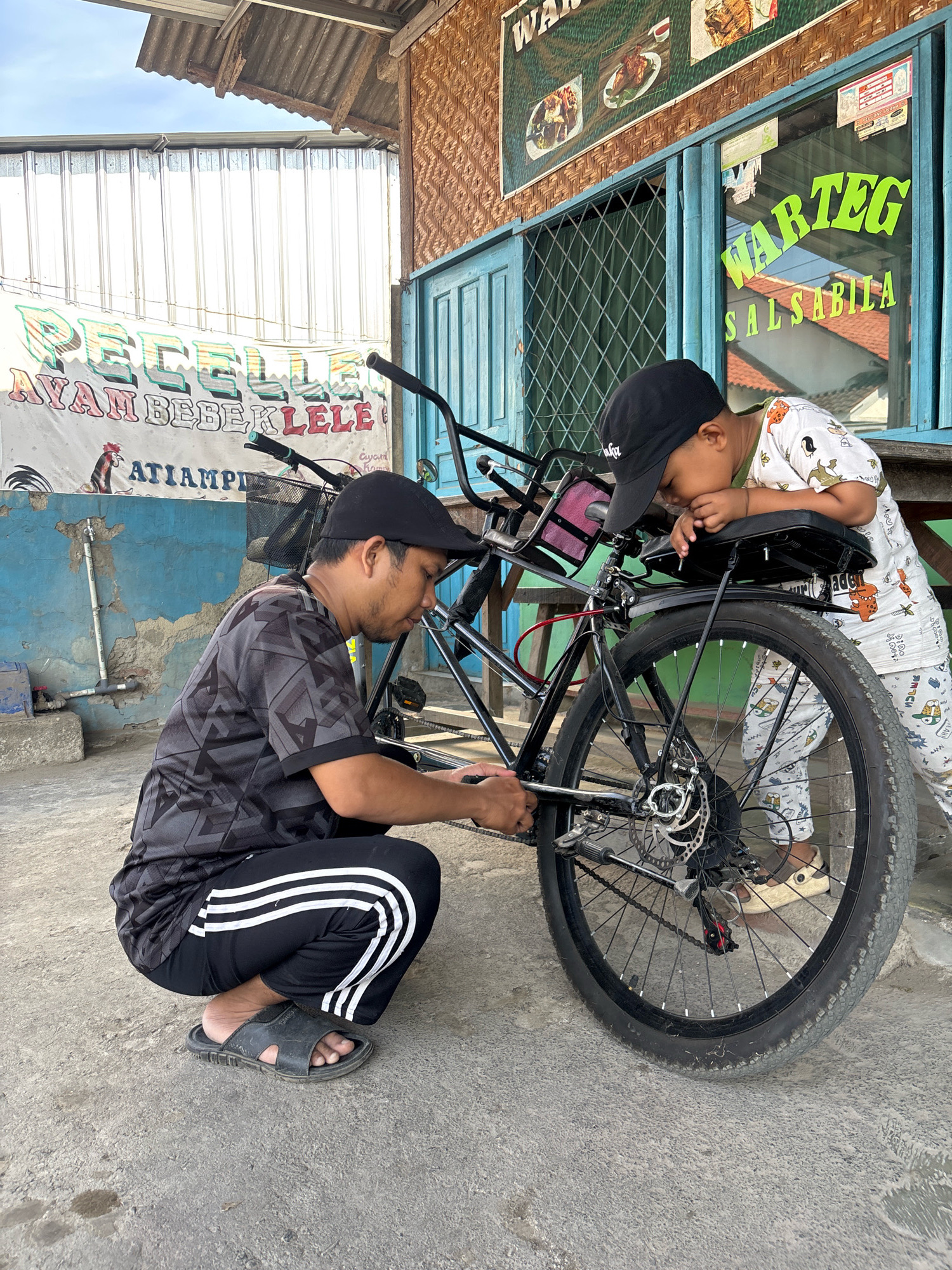 In the city of Tegal, Indonesia, a man kneels and works on the rear wheel of a bicycle while a young boy leans over the bicycle's seat, observing him. The man, wearing a dark patterned t-shirt and black athletic pants with white stripes, is crouched on the concrete ground. The boy, dressed in a white t-shirt with cartoon patterns and light-colored pants, is wearing a black baseball cap. They are situated outside a building with a blue door and window frames, adorned with signage. One sign above the window reads "WARTEG SALSABILA" in bright green lettering, indicating a local eatery. Another banner behind the bicycle displays "PECELL LELE AYAM BEBEK LELE ATIAMPI" in colorful fonts, also suggesting food offerings. The background reveals a corrugated metal roof and walls, typical of Indonesian street-side establishments. The overall scene depicts a moment of shared activity and perhaps a lesson in bicycle maintenance in a casual, urban setting.