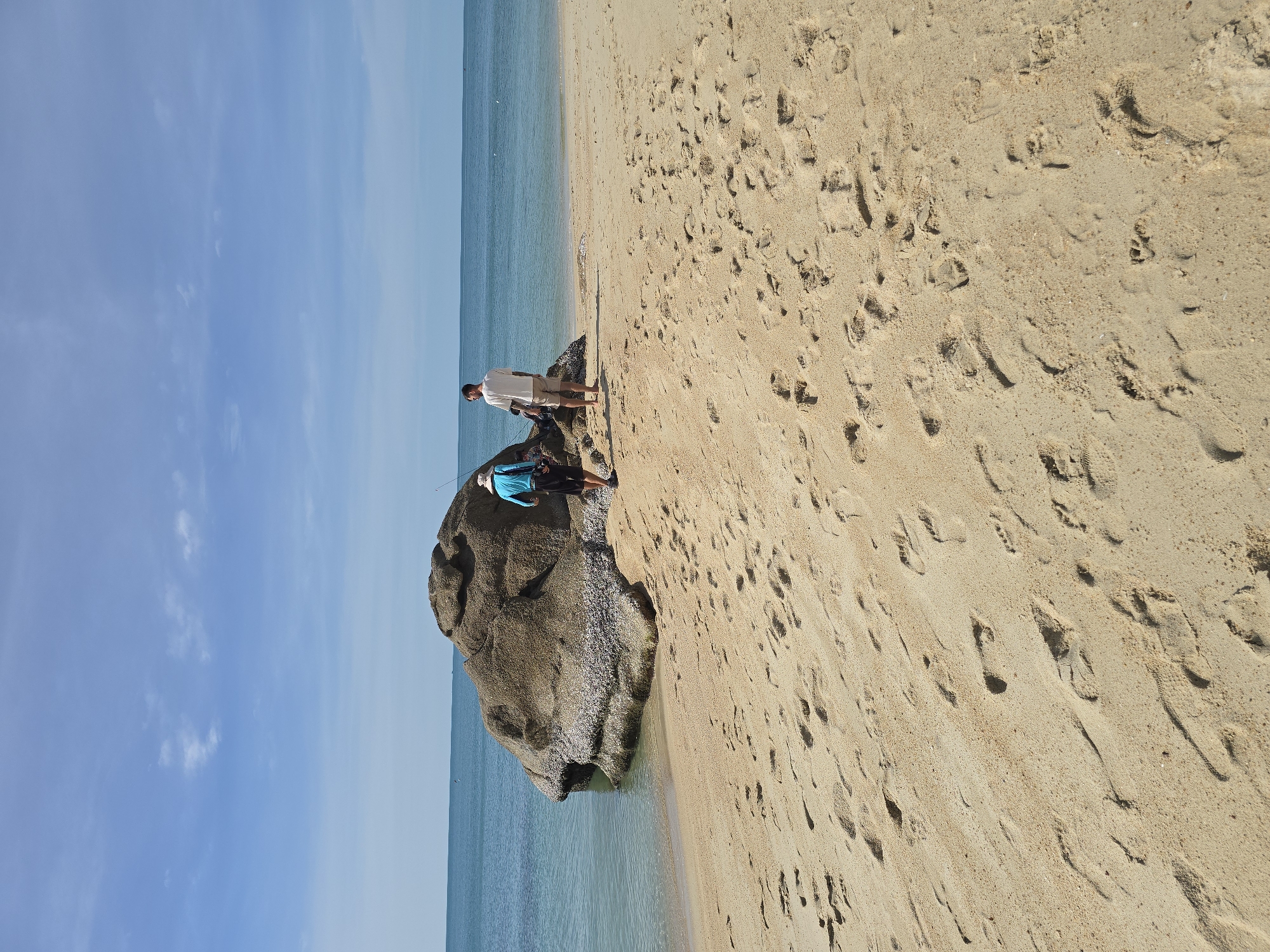 Two individuals stand on a sandy beach with a large rock formation. One person, wearing a blue shirt and hat, walks away from the camera towards the rock. The other person, in a white t-shirt and shorts, stands facing the water. The ocean is calm and blue, meeting a clear sky with scattered clouds. The sand is marked with numerous footprints. The scene is located on the beach in Baan Lamai, Thailand.