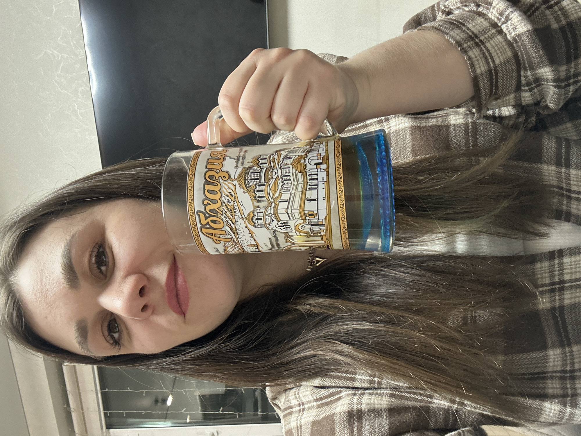 A woman with long brown hair, wearing a brown and white plaid shirt, is holding a clear glass mug in her right hand. The mug features a white and gold image of a building, possibly a cathedral or church, and the Cyrillic word "Ryazan". The mug appears to be partially filled with a dark liquid. The background shows a dark television screen and a window with what appear to be blinds or curtains. The picture is rotated to the side.