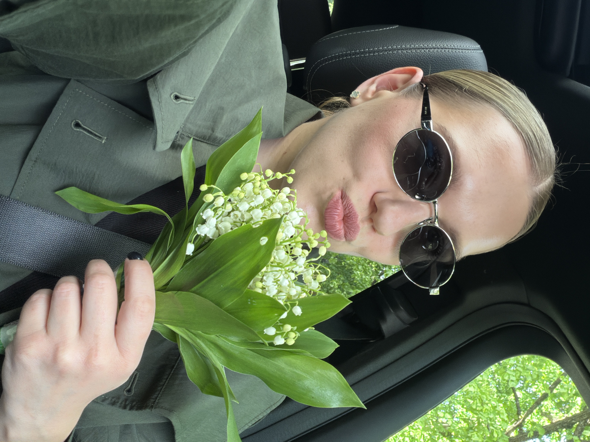 A woman wearing sunglasses is holding a bouquet of lilies of the valley inside a car. She has fair skin and her hair is blonde. She is wearing a green collared shirt. She is in the driver's seat of a car with a black interior. The setting is likely outdoors given the presence of a tree and green foliage seen through the car window. However, there is no information about the city of Riga, Latvia, or anything that might suggest the location.