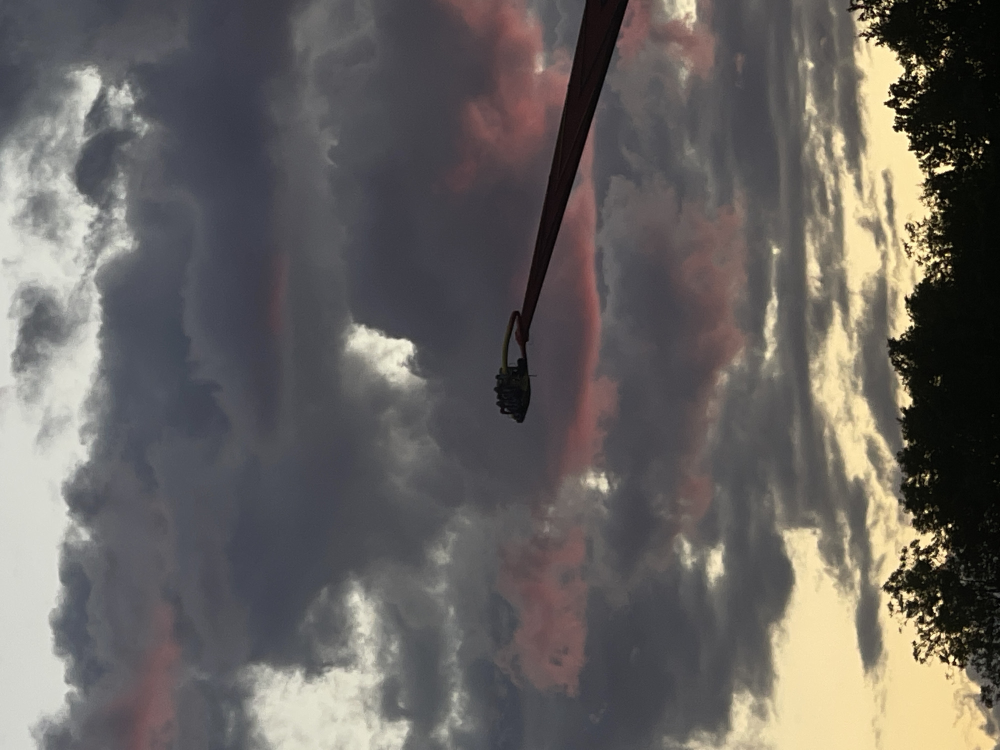 An amusement park ride, possibly a swing ride, is visible against a dramatic sky filled with dark, ominous clouds tinged with pink and orange. The ride is suspended from a long, red arm, with several gondolas or seats clustered at the end of the arm. Below the sky, silhouetted trees form a dark line across the bottom of the frame, suggesting the ride is located in a park or outdoor recreational area. The lighting indicates it is likely dusk or dawn, given the interplay of light and shadow in the clouds and the silhouette of the trees. There are no people clearly visible on the ride. The overall impression is one of dramatic natural light interacting with man-made amusement park structures.