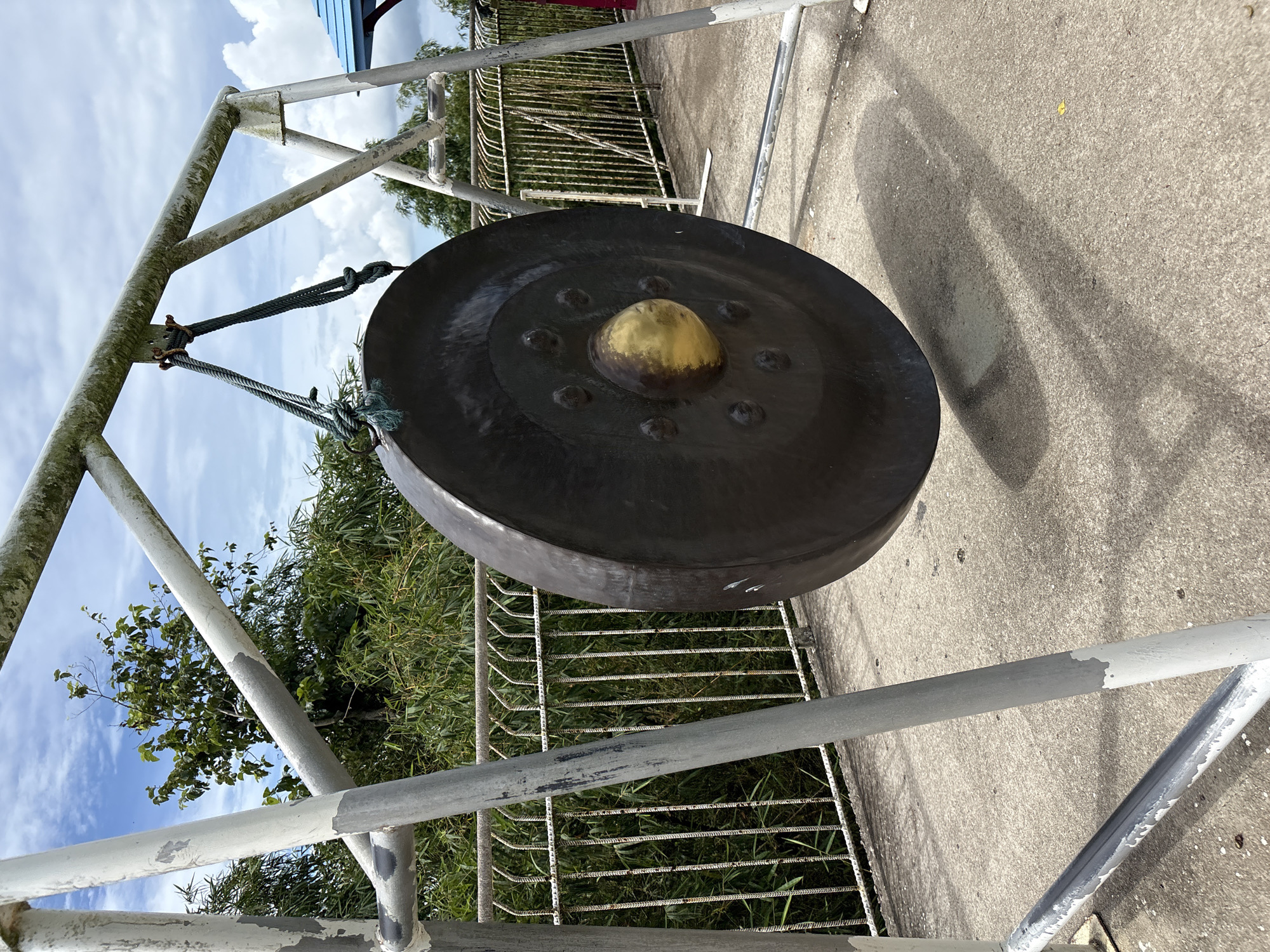 A large, dark-colored gong with a golden, raised center hangs from a rusty metal frame. The frame is painted white, but the paint is chipped and peeling, revealing rust underneath. The gong is suspended by thick, dark green ropes tied to a metal hook. The scene is outdoors, with a concrete ground and a metal railing in the background. Beyond the railing, there are lush green trees and bamboo. The sky above is cloudy, with patches of blue. The gong casts a long shadow on the concrete. In the far background, a portion of a blue and red structure is visible. This image was taken in Rawai, Thailand.