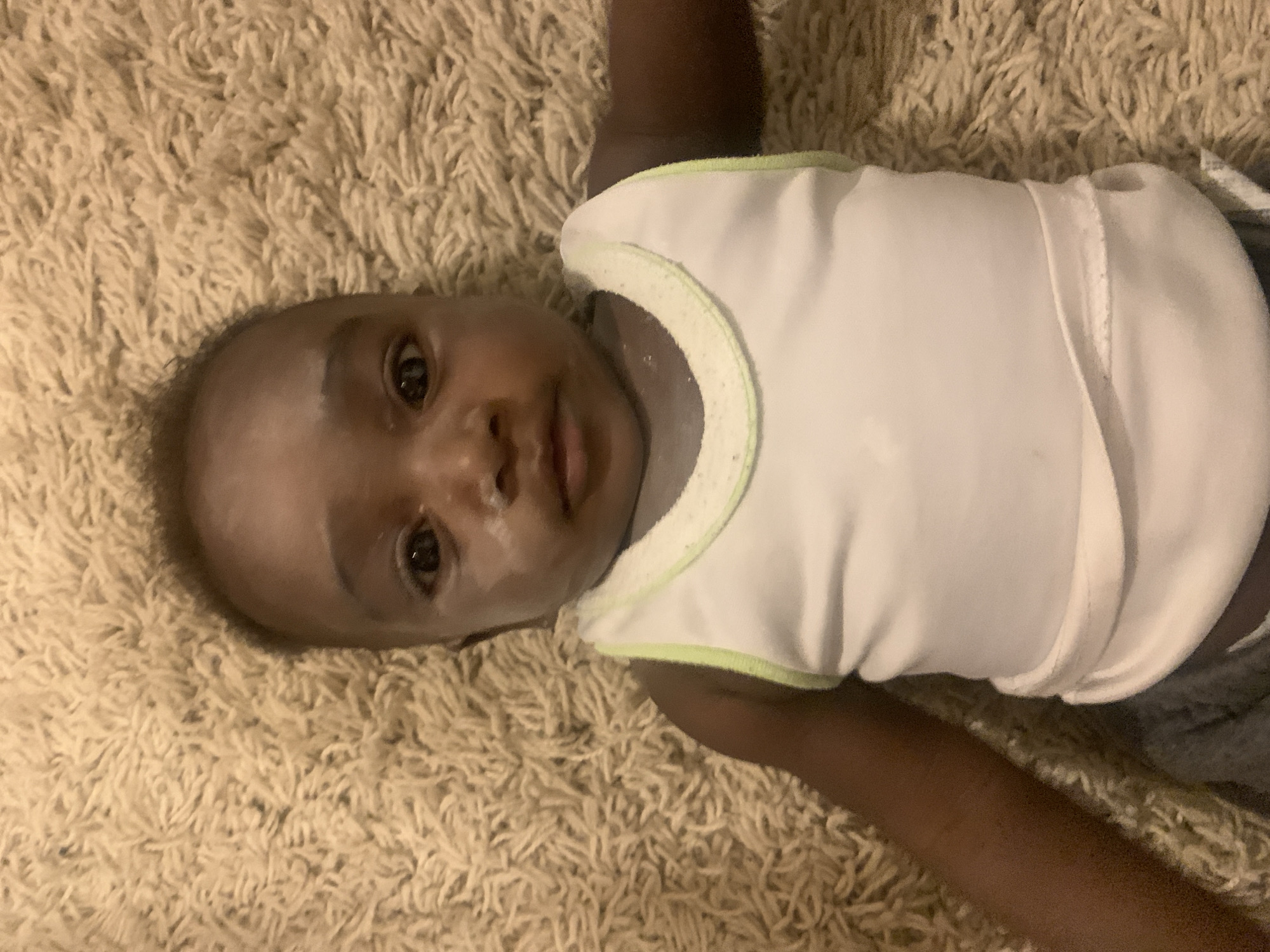 A medium shot captures a baby lying on their back on a light beige, shaggy carpet. The image is rotated, presenting the baby with their head towards the left side of the frame. The baby has dark skin, wide dark eyes, and a neutral, curious expression, looking directly at the camera. They are wearing a plain white sleeveless undershirt with a distinctive light green trim around the neck and armholes. White residue, possibly baby powder or dried liquid, is visible on the baby's face, particularly around the nose and cheeks.
The background is dominated by the textured, plush carpet. A small portion of what appears to be grey fabric, possibly a blanket or part of another garment, is visible near the baby's lower right side. The scene is brightly lit, consistent with an indoor setting, and no specific time of day is discernible. There is no visible text in the image. The baby appears calm and observant, lying still on the soft surface. This moment likely takes place in a home environment in Barakin Akawo, Nigeria.